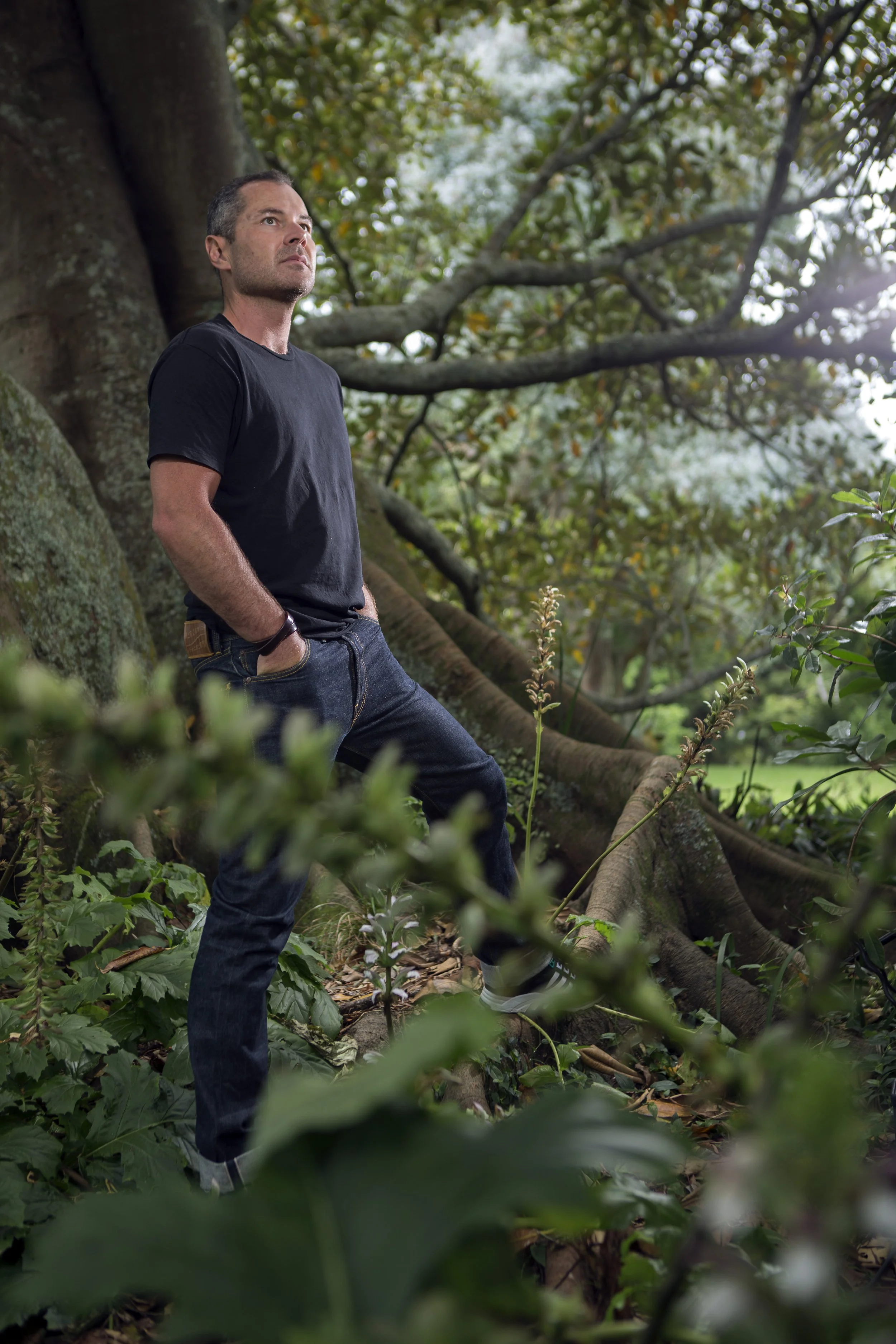 A man standing outdoors next to a large tree in a wooded area, leaning against the trunk with hands in pockets, looking contemplative.