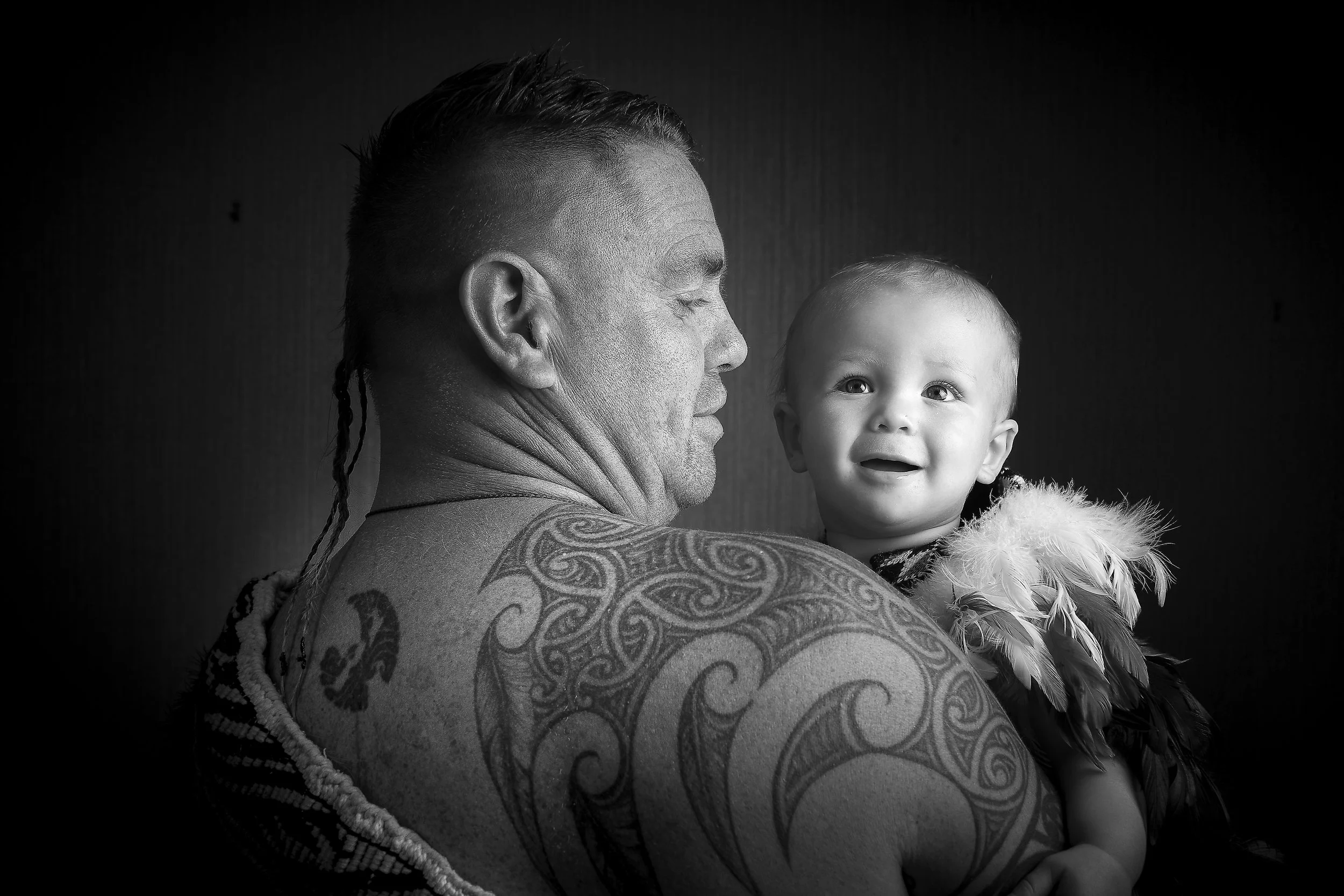 Black and white photo of a man holding a young child, both facing each other. The man has tattoos on his shoulder and back, and the child appears to be crying or upset.