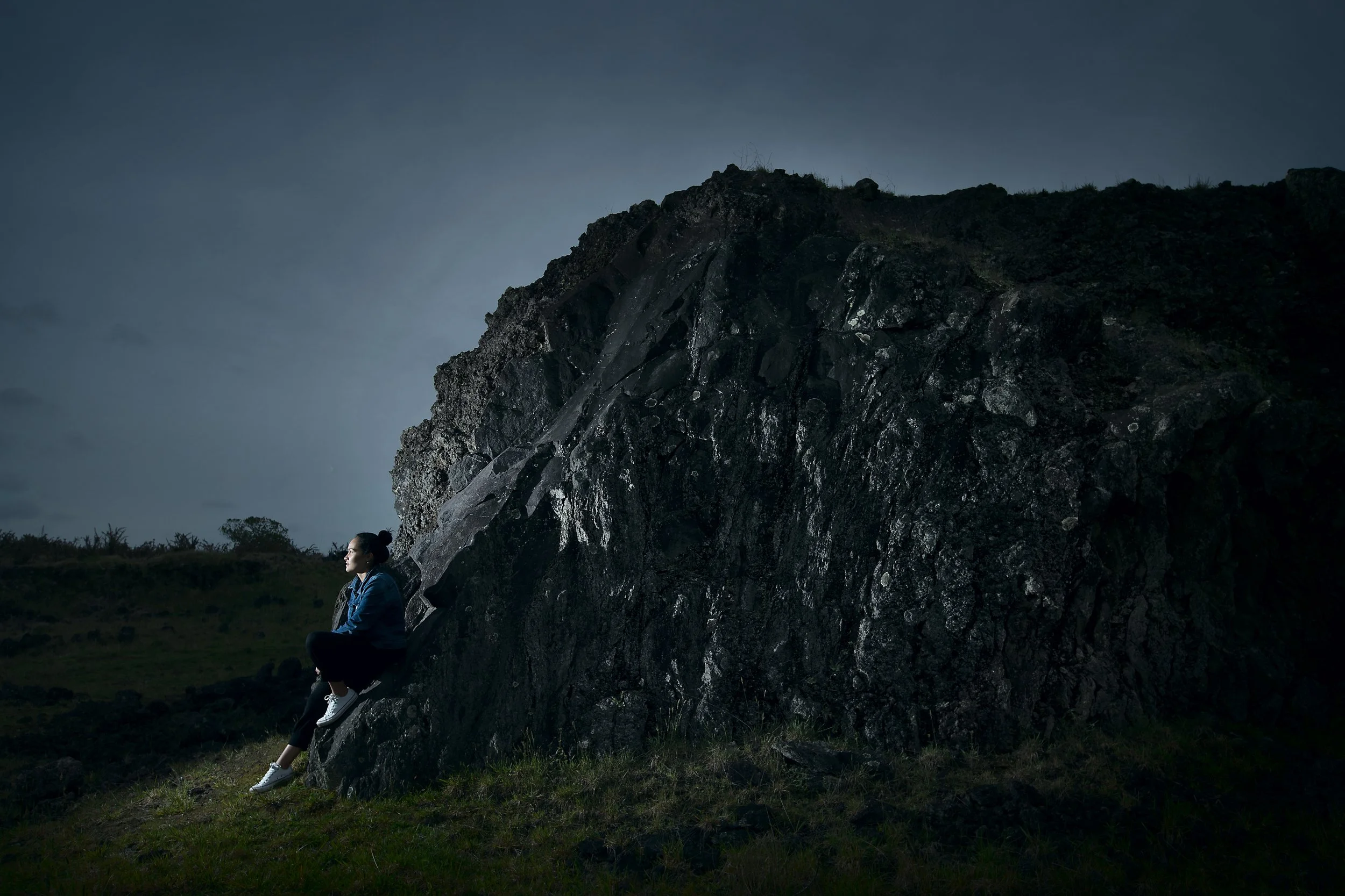 A person sitting on a rock at dusk, looking thoughtful, with a large dark rock formation and cloudy sky in the background.