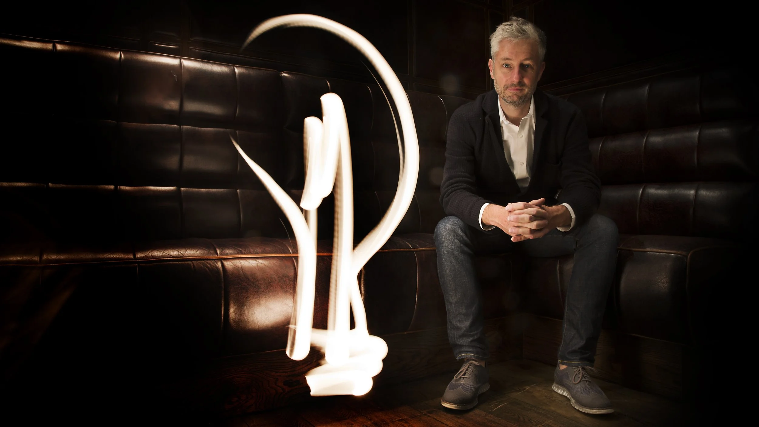 A man sitting on a dark leather banquette in a dimly lit room with a modern, abstract light sculpture beside him.