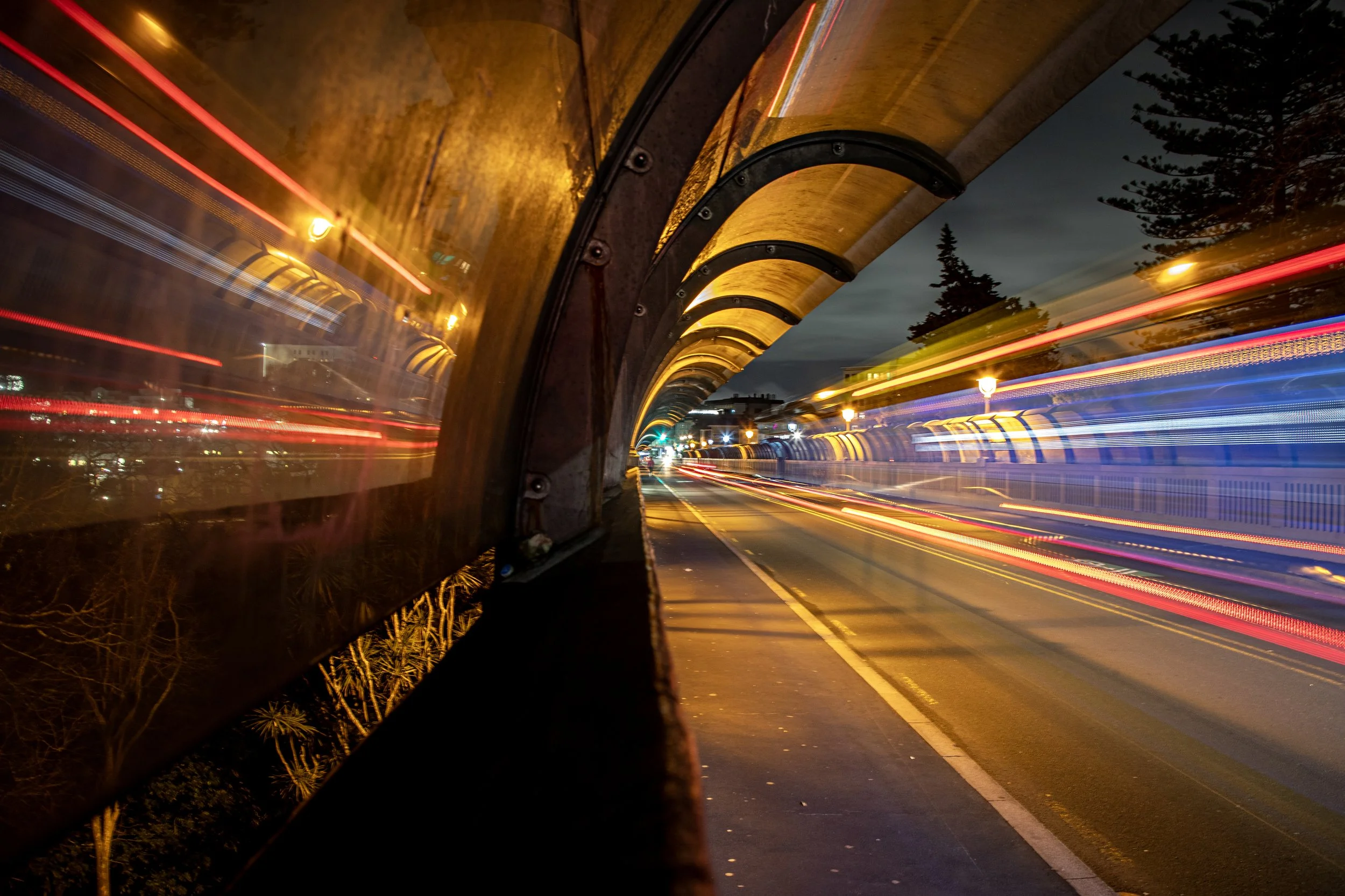 Nighttime street scene viewed through a bus window with a yellow canopy, showing light trails from passing vehicles and streetlights.