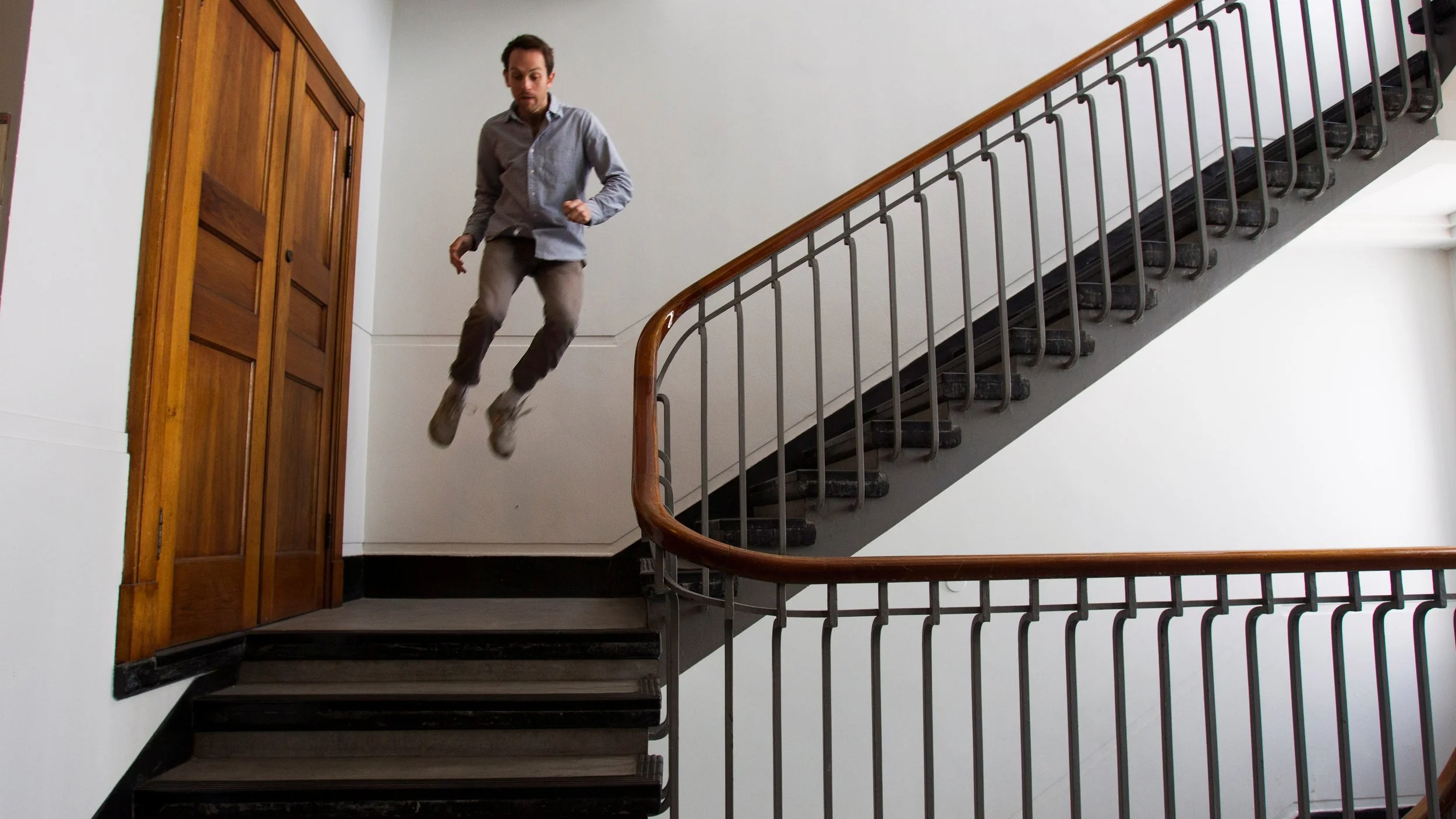 A man mid-air as he jumps down a staircase, with a railing and wooden door nearby.