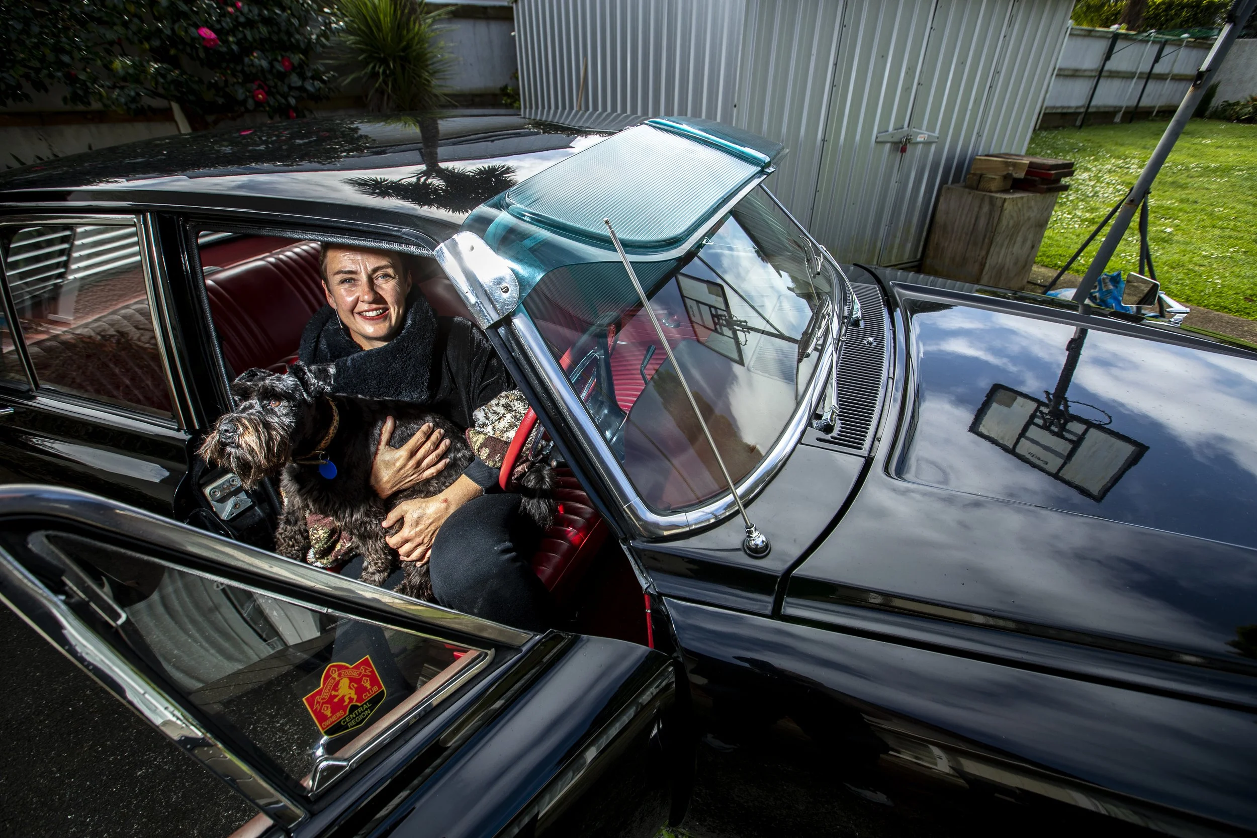 A woman sitting inside a vintage black car, smiling and holding a dog on her lap, with the car's roof partially open. The interior has red seats, and the background shows a yard with greenery, a basketball hoop, and a metal fence.