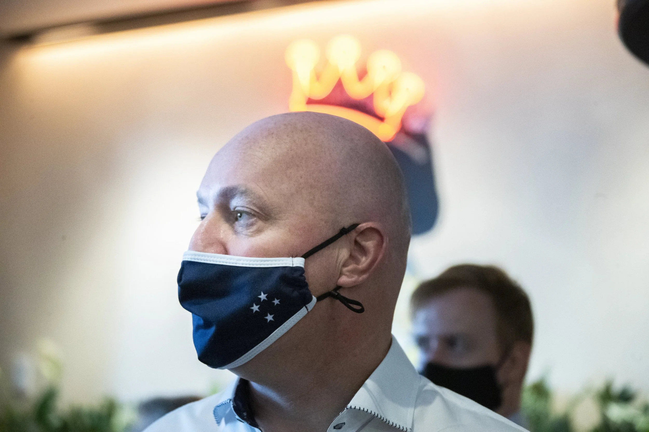 A bald man with a facial mask featuring stars, looking to the side, with another person wearing a black mask in the background. A glowing crown-shaped sign is in the background.
