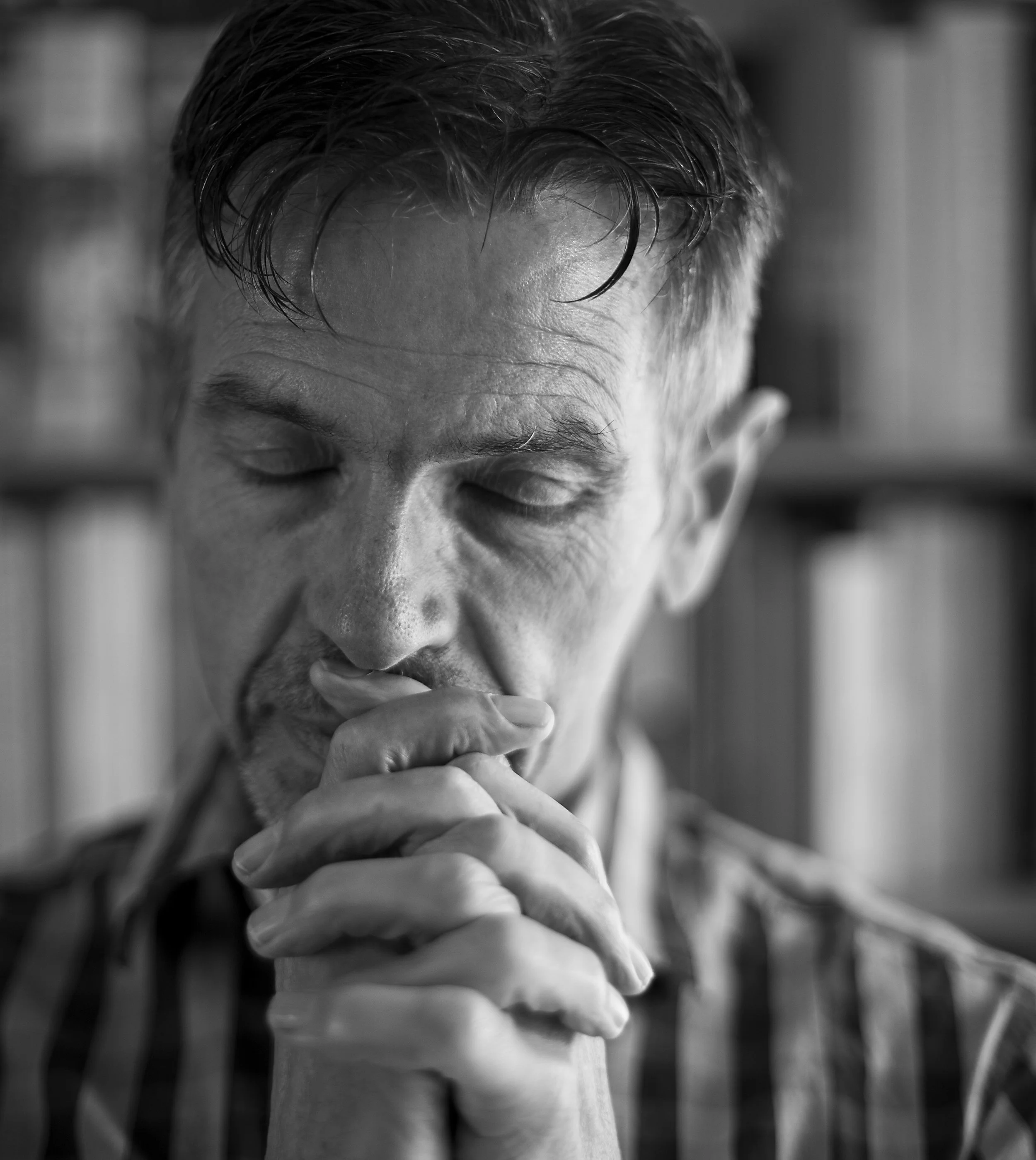 Black and white close-up of a man with closed eyes, resting his chin on his clasped hands appearing contemplative.