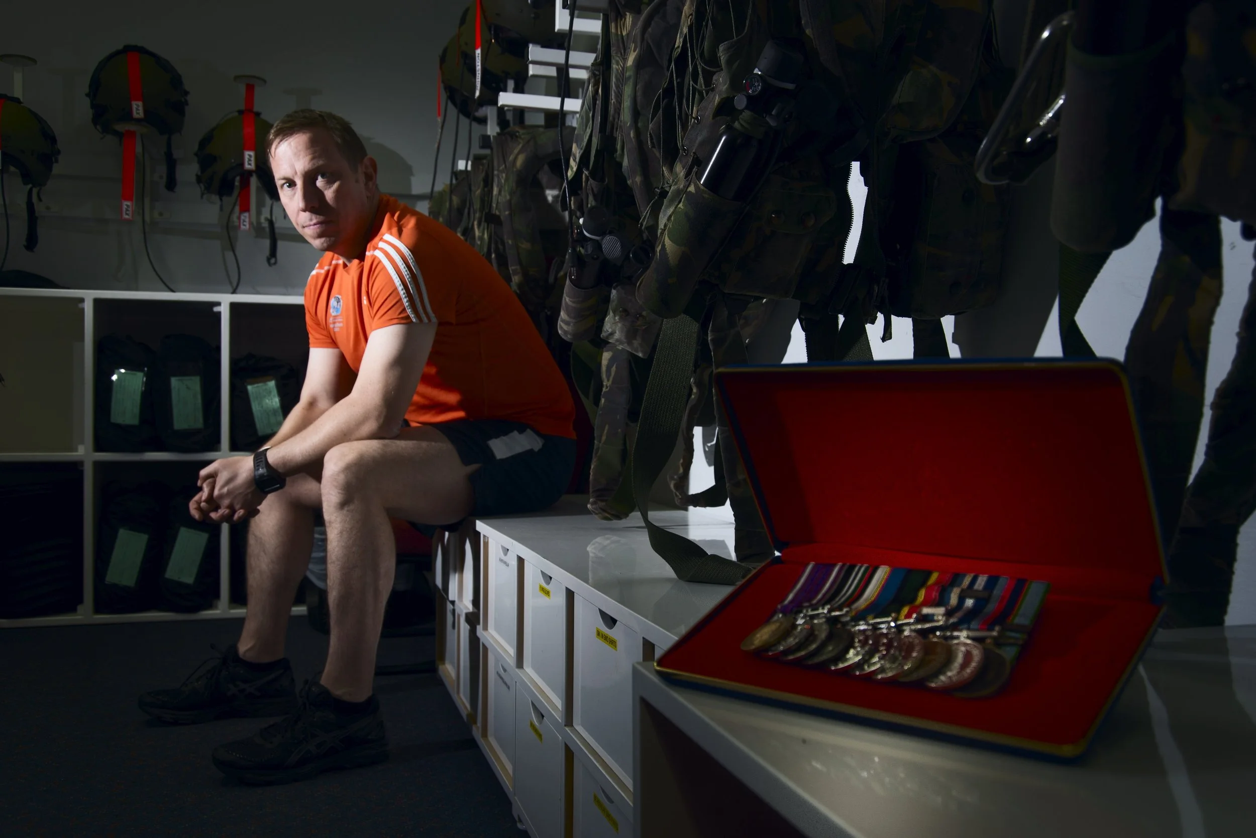 A man sitting on a bench in military gear storage room, with medals in an open case on a table nearby.