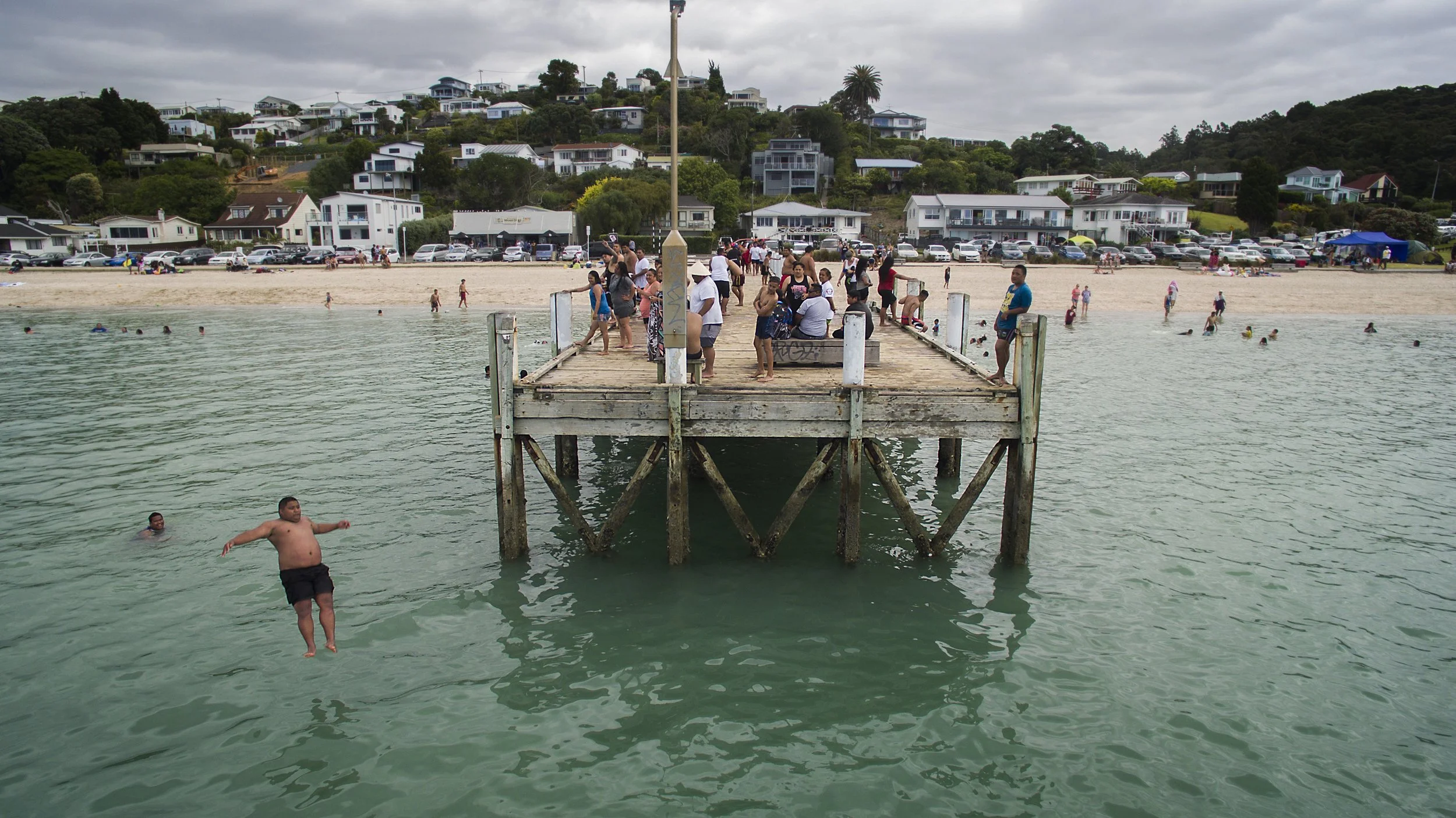 People gathering on a wooden pier extending into a harbor with swimmers in the water, against a background of beachfront houses and an overcast sky.
