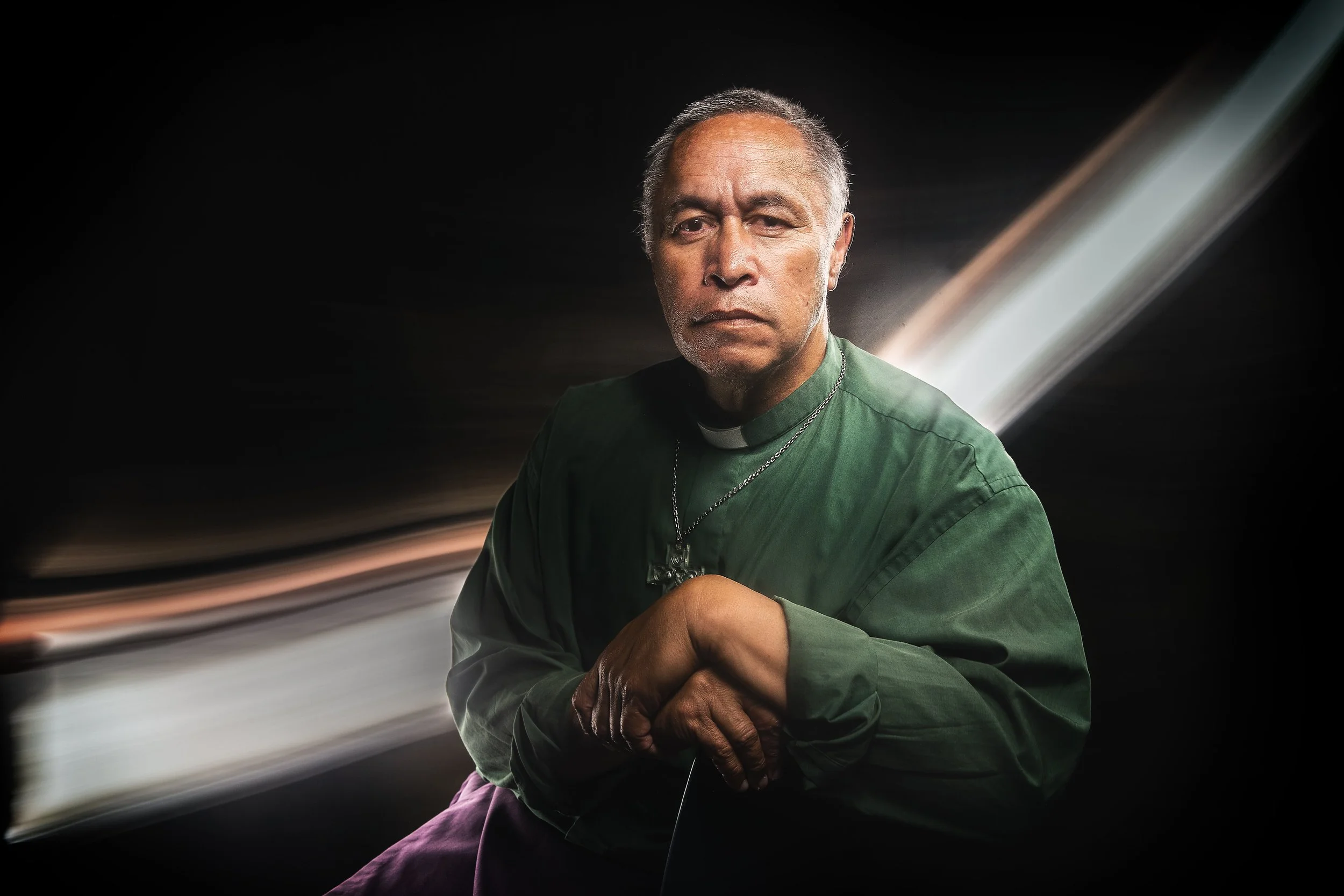 A man dressed as a priest in a green robe with a white clerical collar and a cross necklace, looking serious, with a dark background and streaks of light behind him.