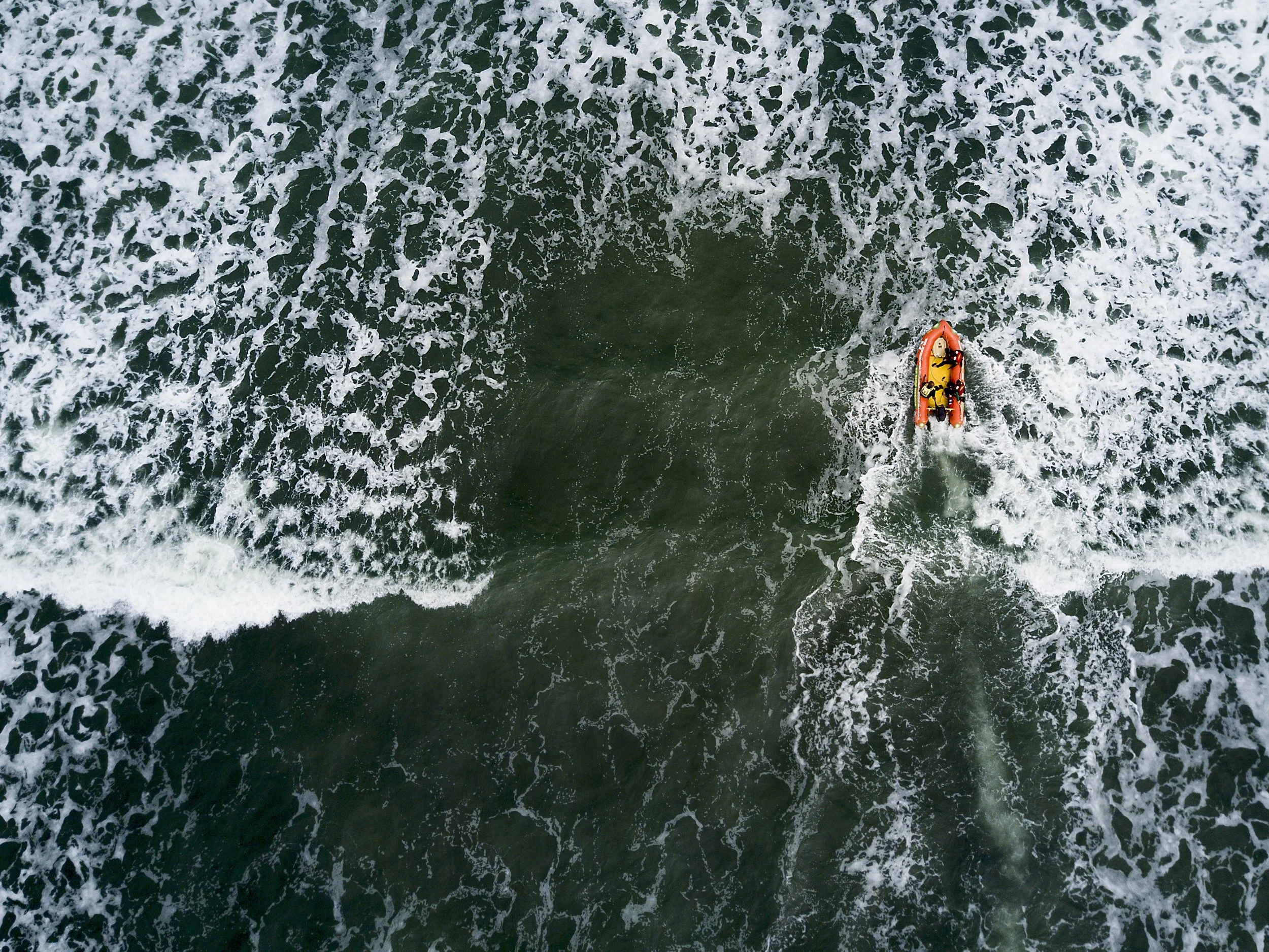 An aerial view of a yellow and orange inflatable boat with three people on it, moving through the water, surrounded by white foamy waves.