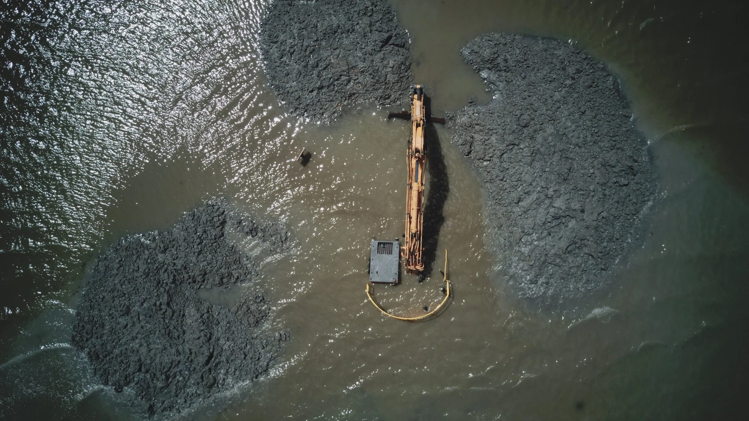 An aerial view of a construction excavator working underwater with piles of mud or dirt on either side in a body of water.