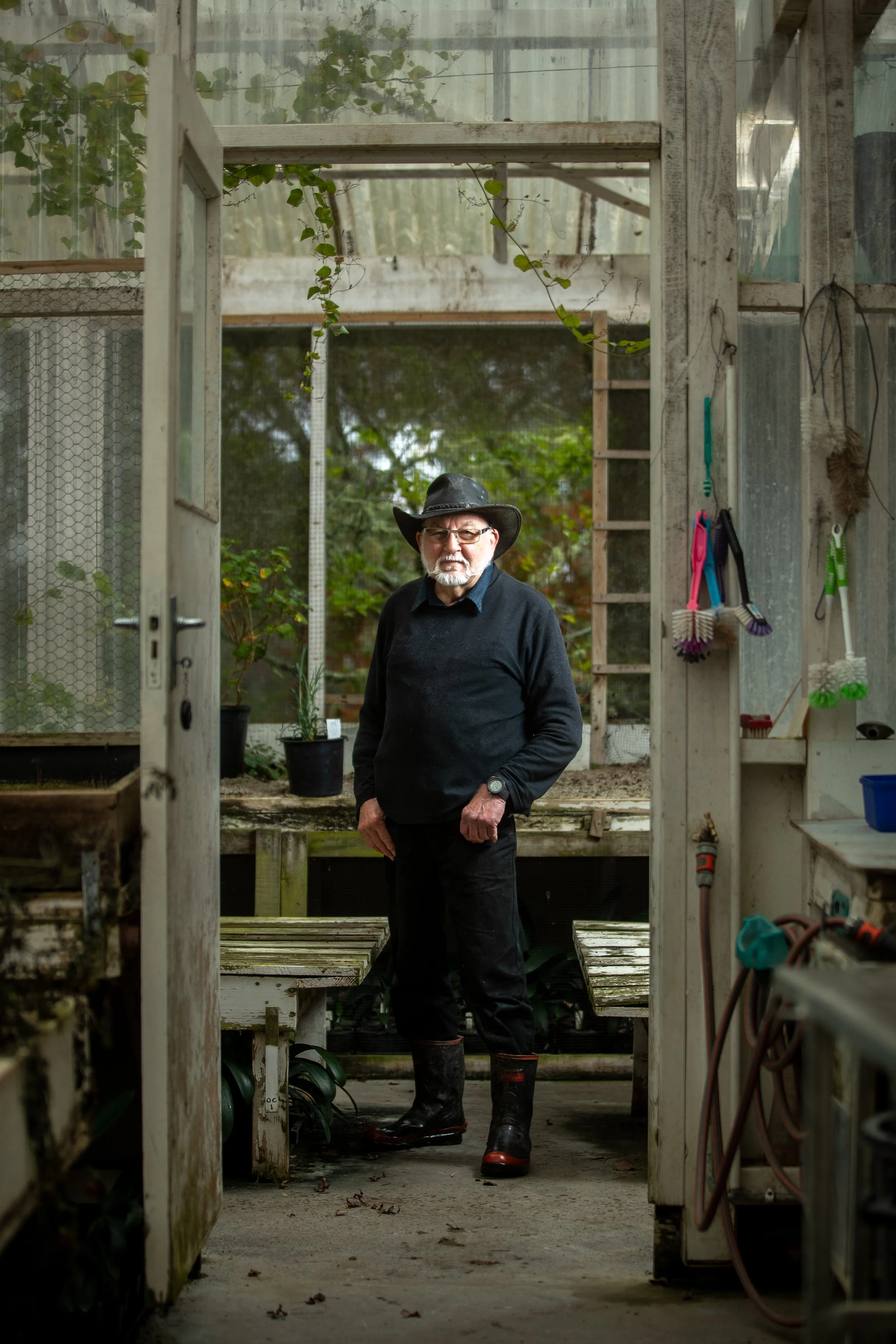 An older man with glasses, a black hat, black sweater, black pants, and rubber boots standing inside a greenhouse or garden shed with plants and gardening tools around him.