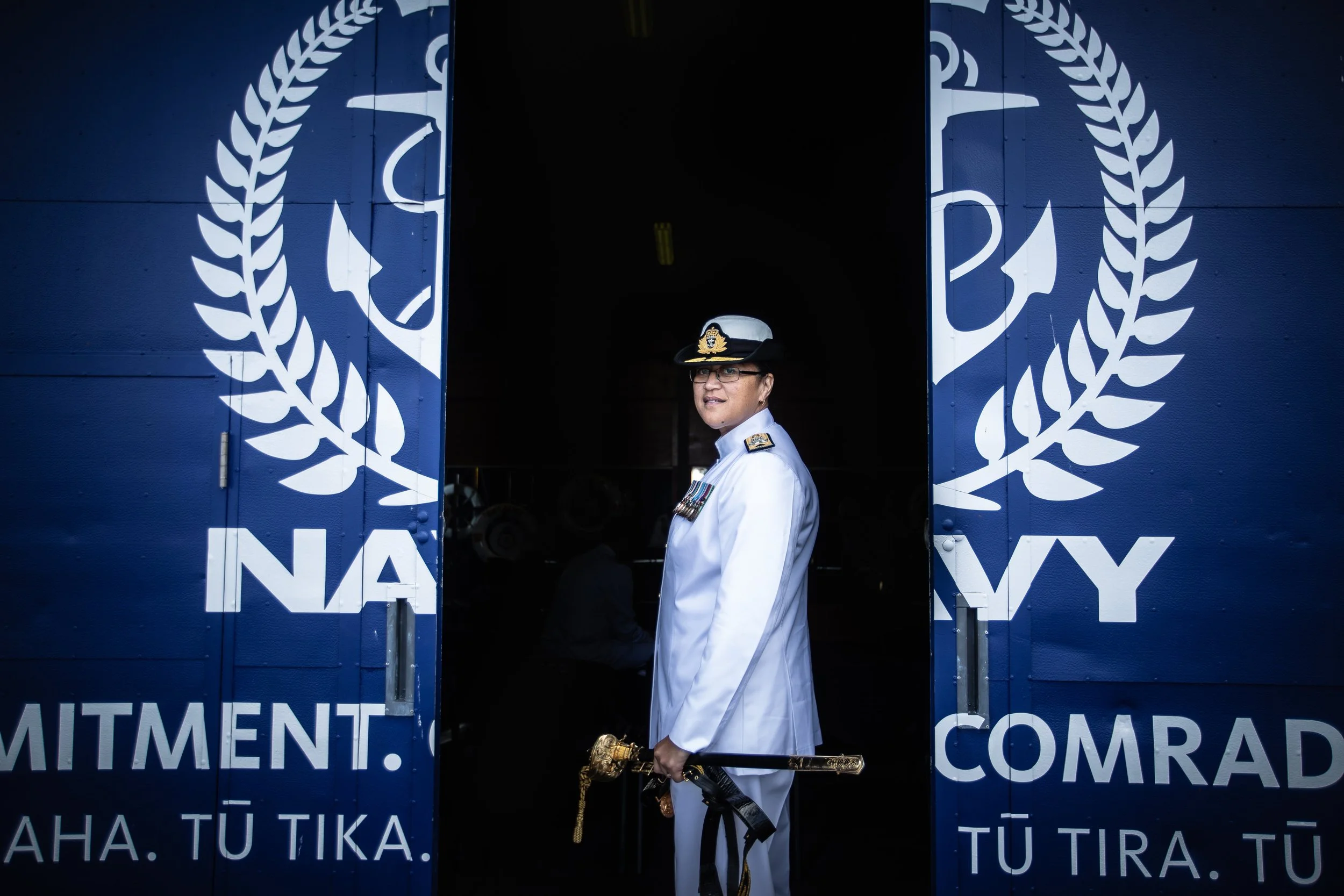 A navy officer in white uniform and cap standing in a doorway with navy insignia, holding a ceremonial sword.