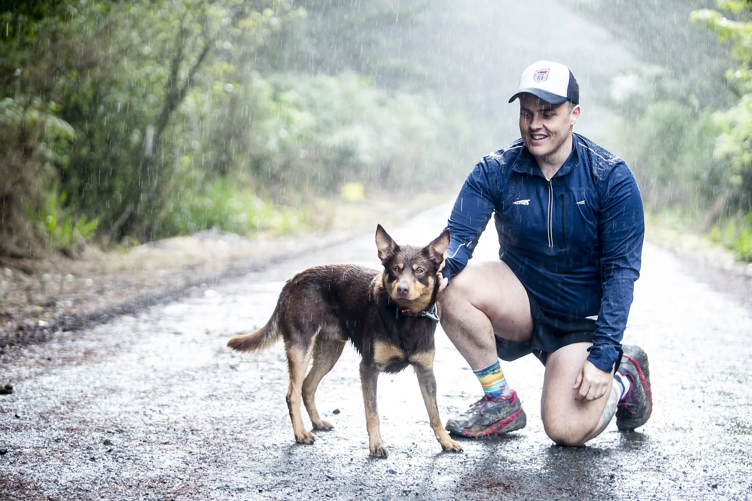 A man kneeling on a wet forest trail holding a dog on a rainy day.
