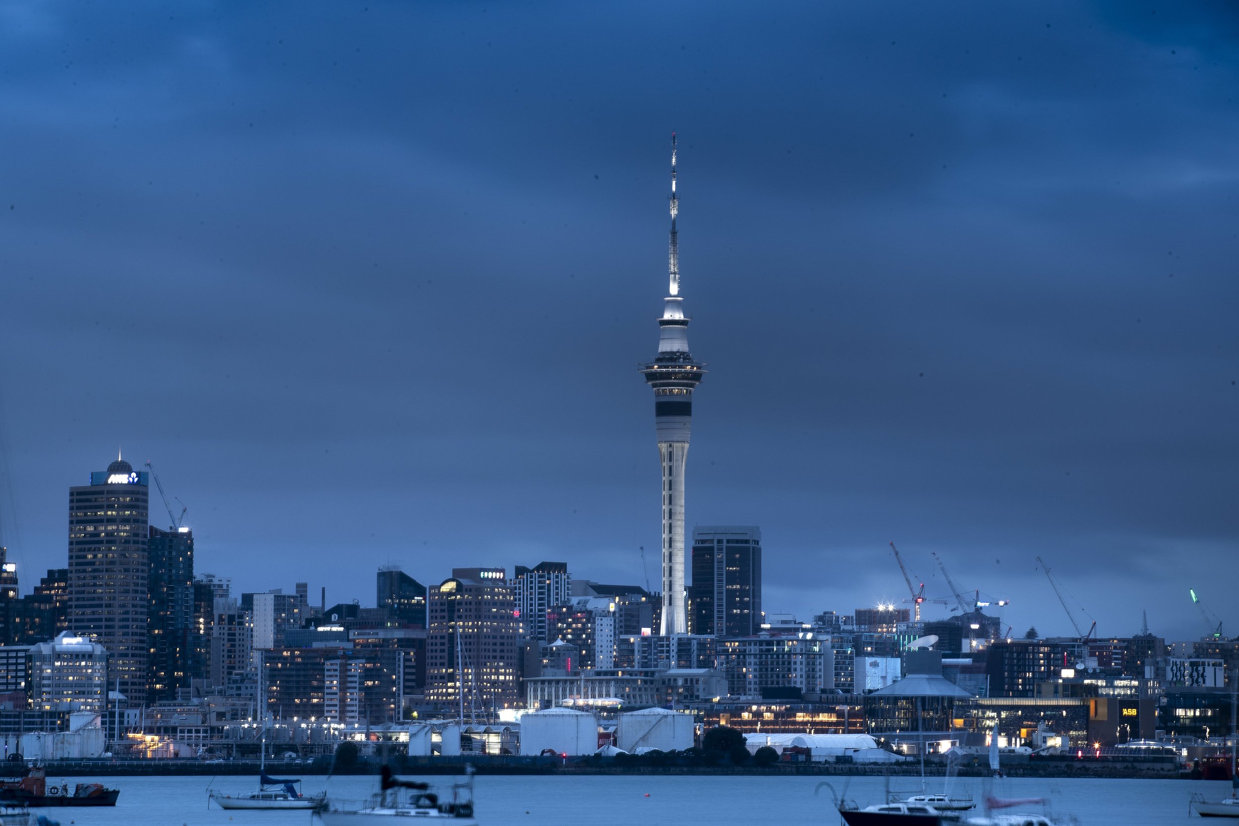 Night view of the city skyline with Auckland Tower in the center, illuminated buildings, a waterfront with boats, and dark cloudy sky.
