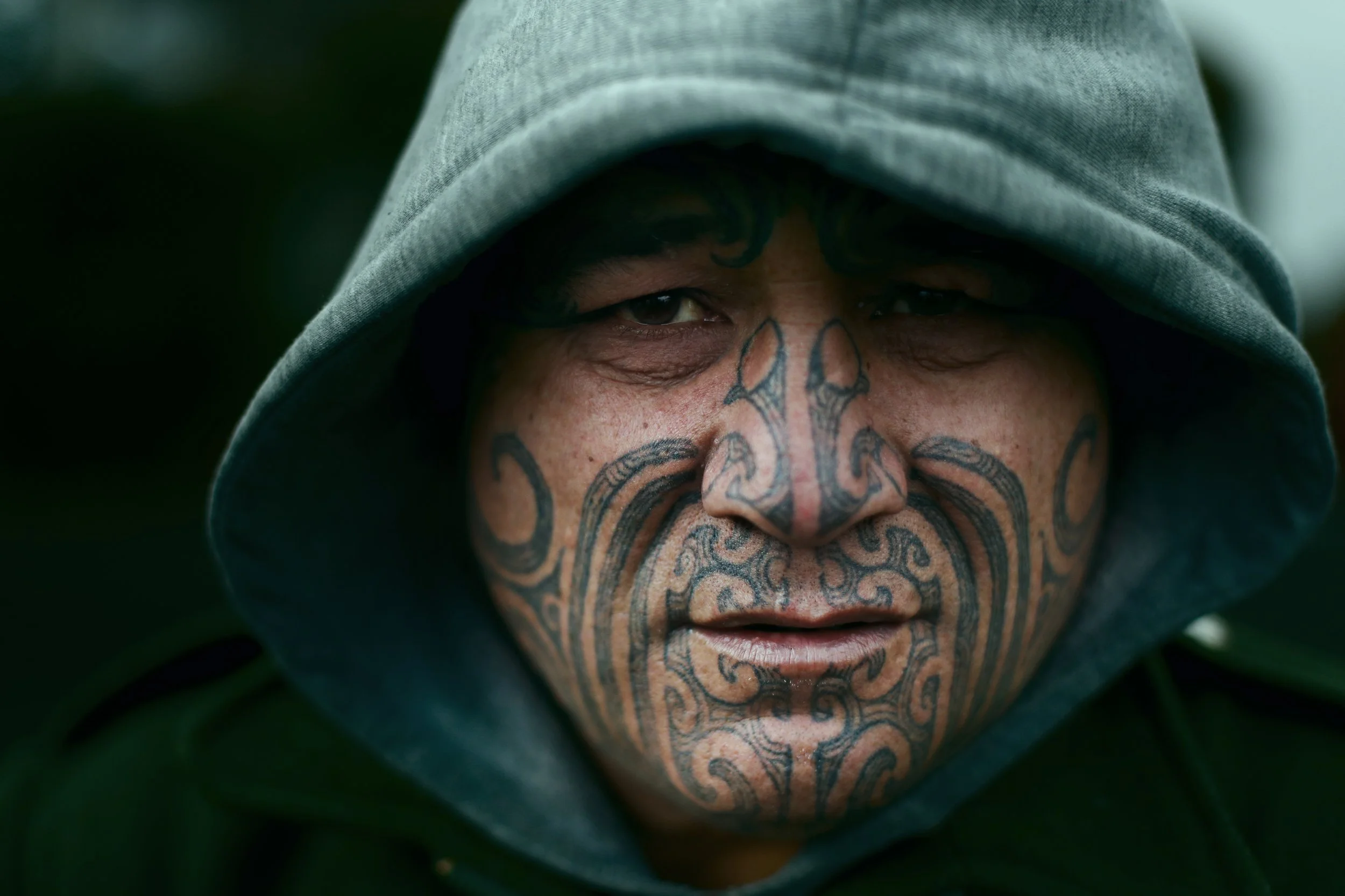 A person wearing a gray hoodie with a tattooed face, featuring intricate black ink designs covering the nose, cheeks, and chin, with partially visible eyes.