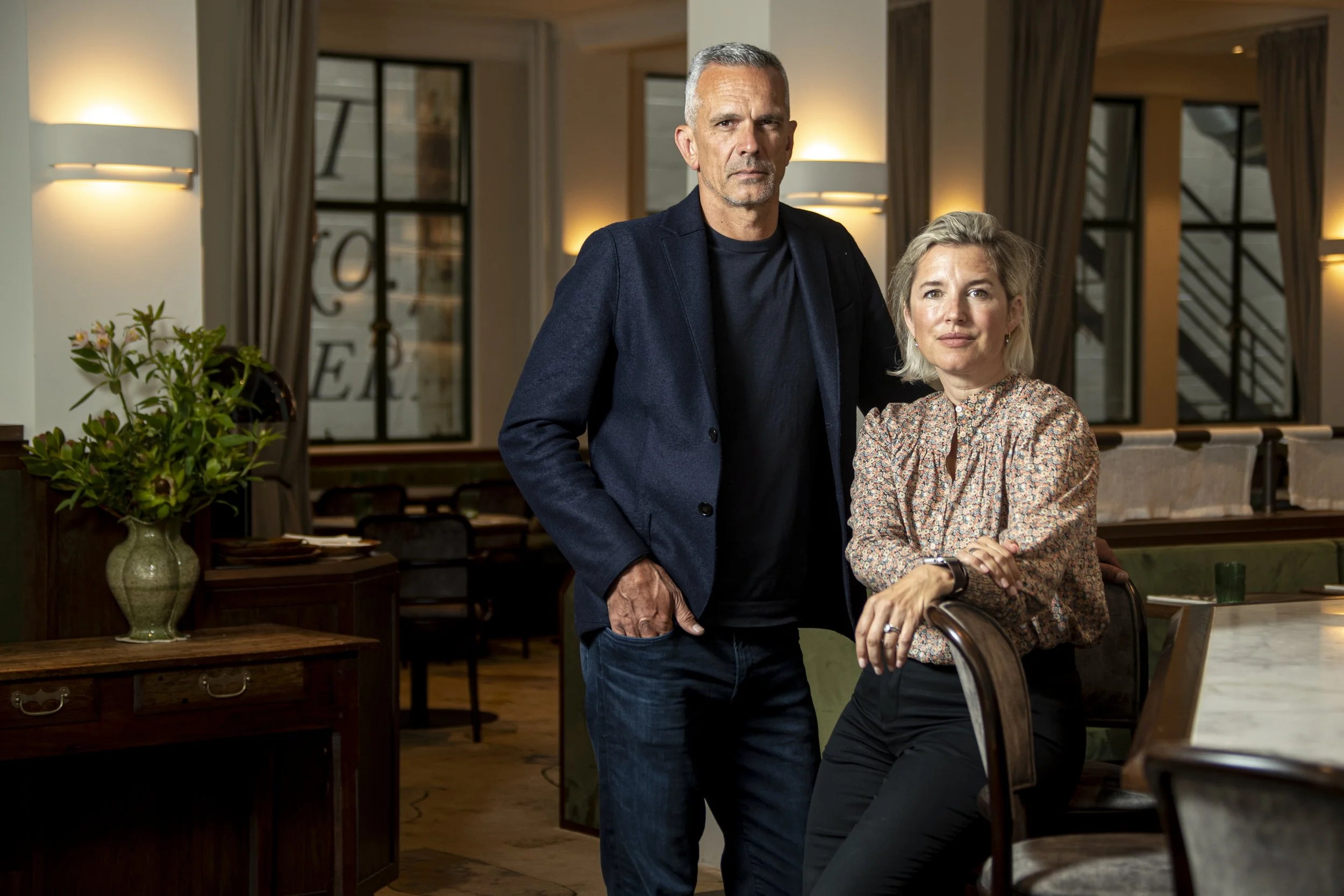 A man with gray hair in a dark blazer standing and a woman with short blonde hair in a patterned blouse sitting in a cozy restaurant or cafe with warm lighting, wooden furniture, and large windows.