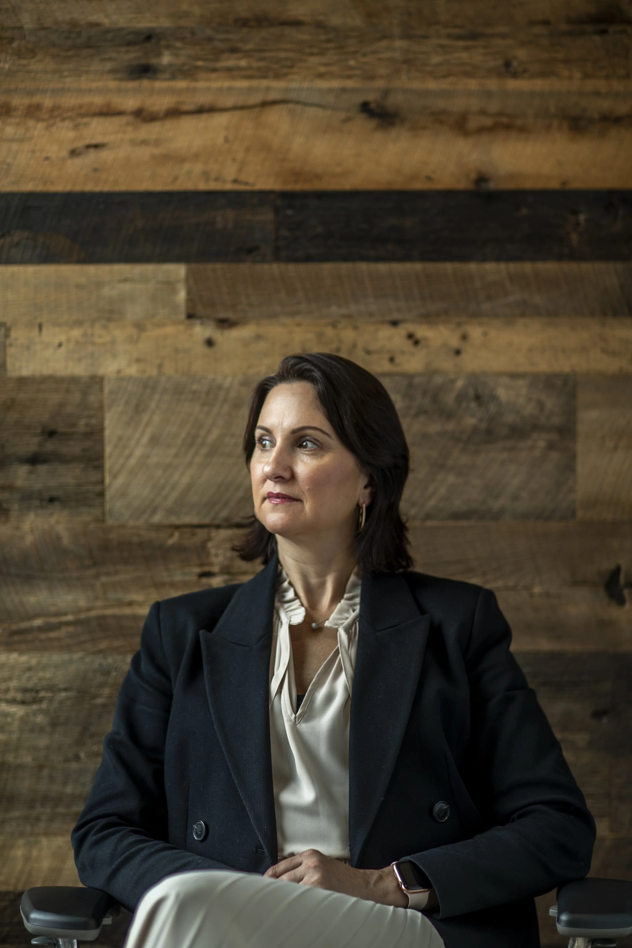A woman sits in front of a wooden wall, dressed in a black blazer and a cream blouse, looking to her right.