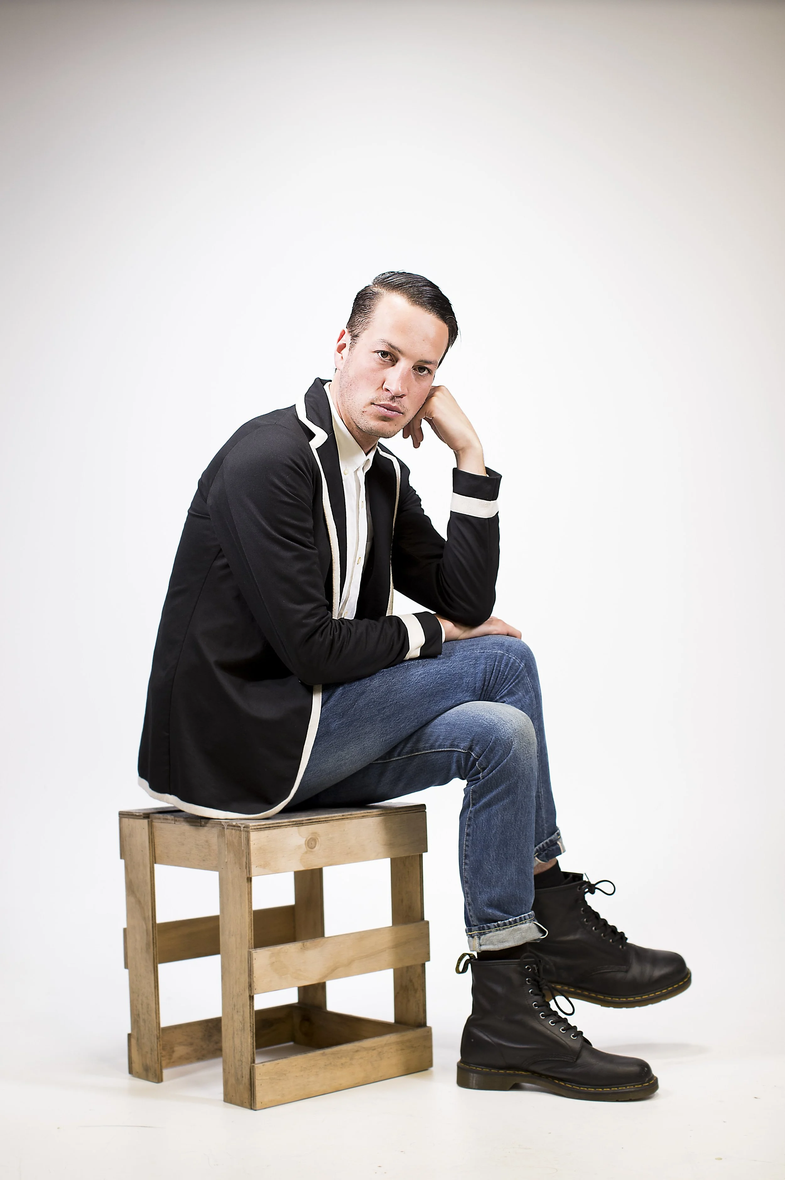 A young man sitting on a small wooden stool against a plain white background, wearing black boots, blue jeans, and a black jacket with white trim, looking at the camera with a neutral expression.