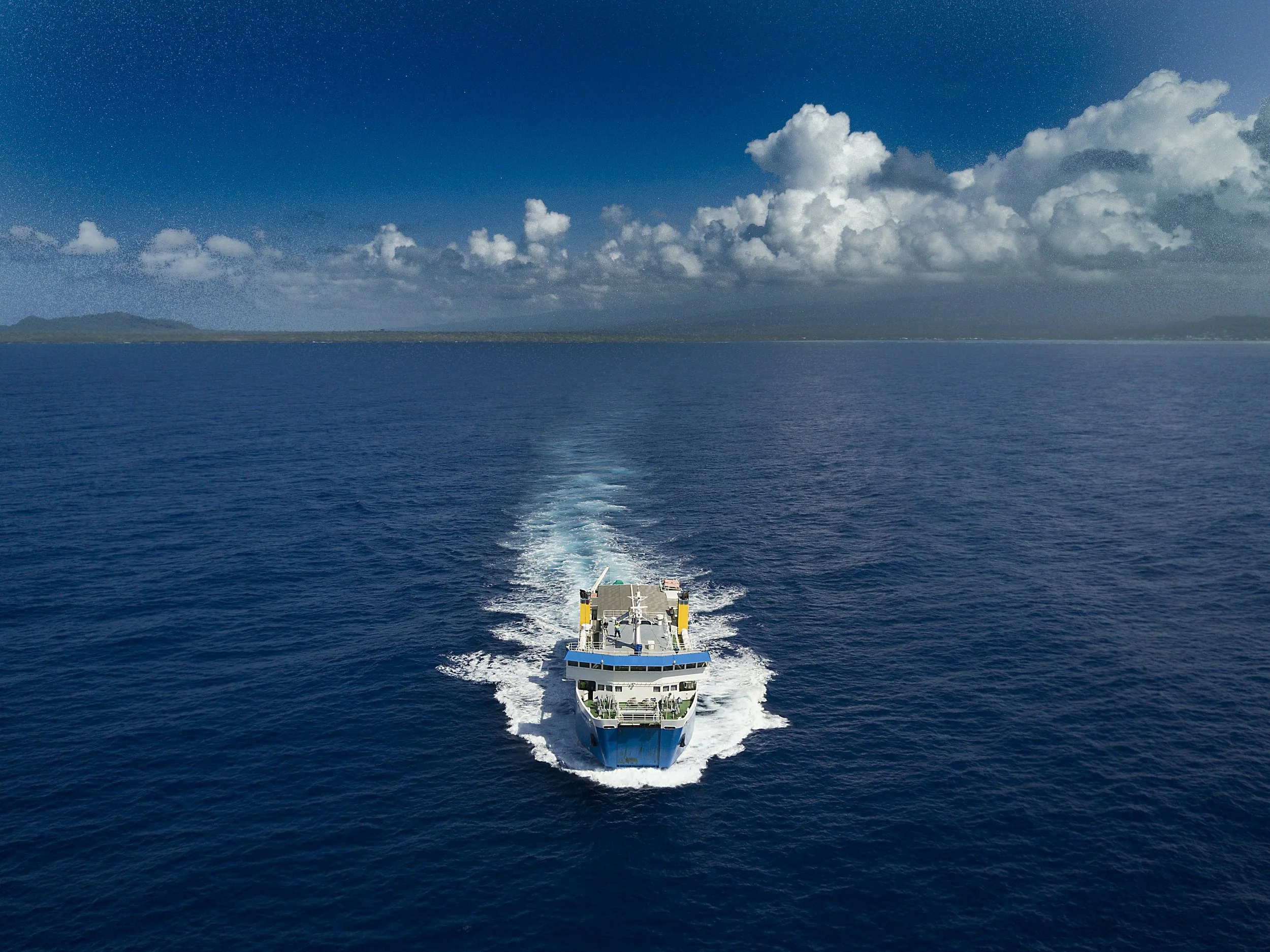 A boat cruising through the ocean, leaving a trail of white waves behind, with a distant island and blue sky with scattered clouds in the background.
