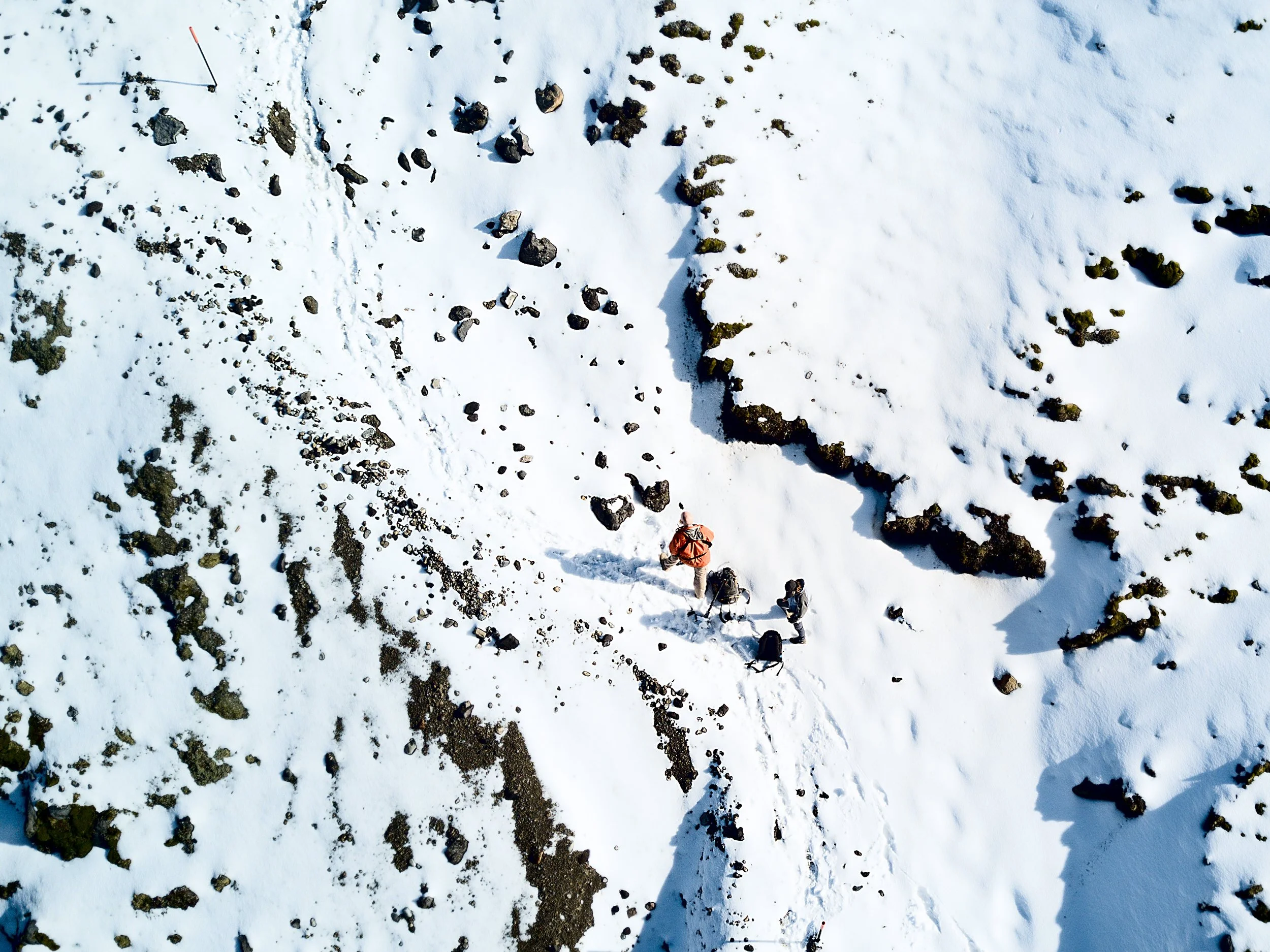 Aerial view of three hikers walking on snow-covered mountain trail with rocks and snow drifts.