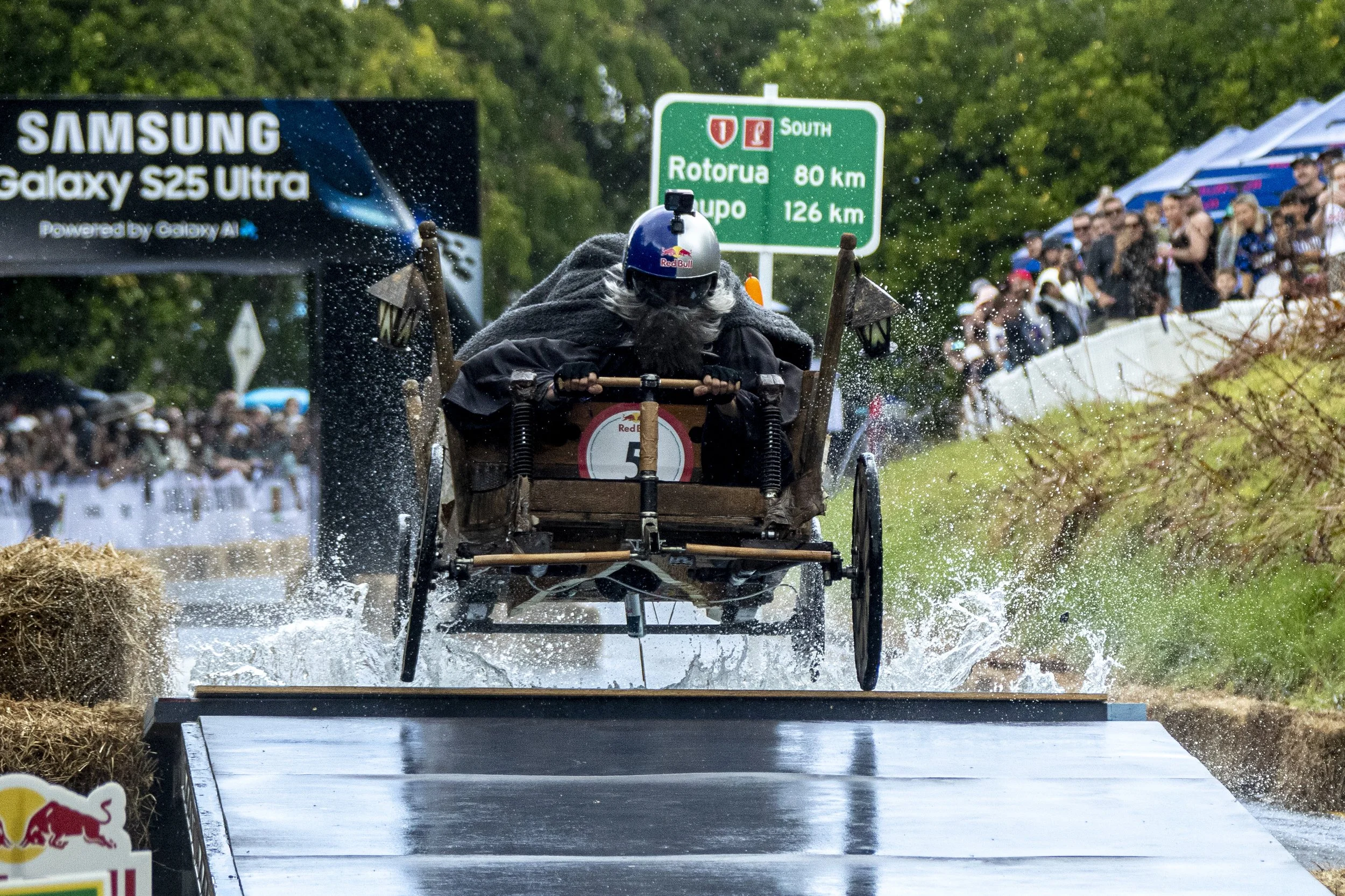 A person riding a wooden cart with large wheels crossing a wet water track, with a crowd of spectators on both sides and greenery in the background.