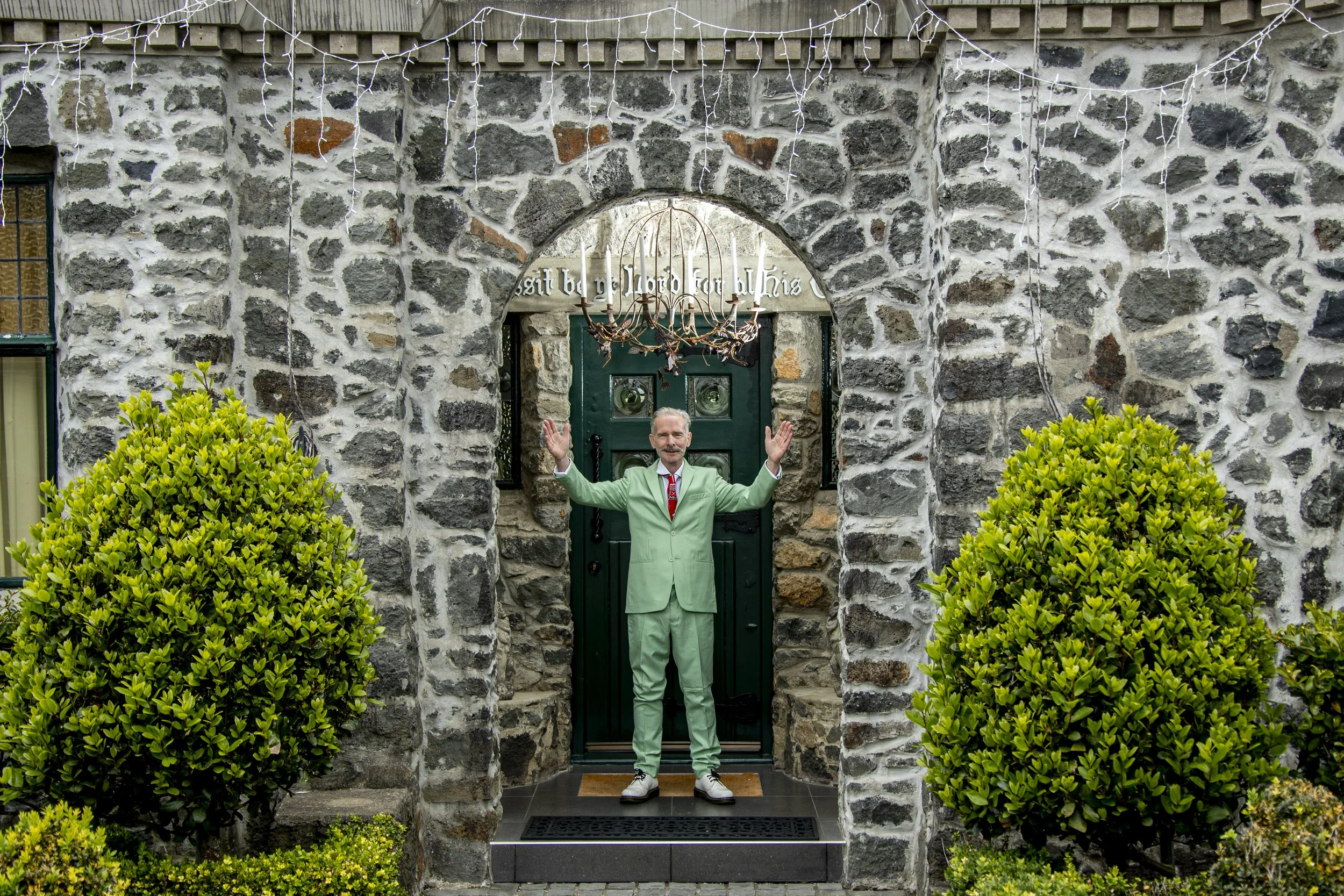 A man in a light green suit standing at a stone house door with arms raised, surrounded by green bushes, with a chandelier hanging above the door, and decorative text above it.