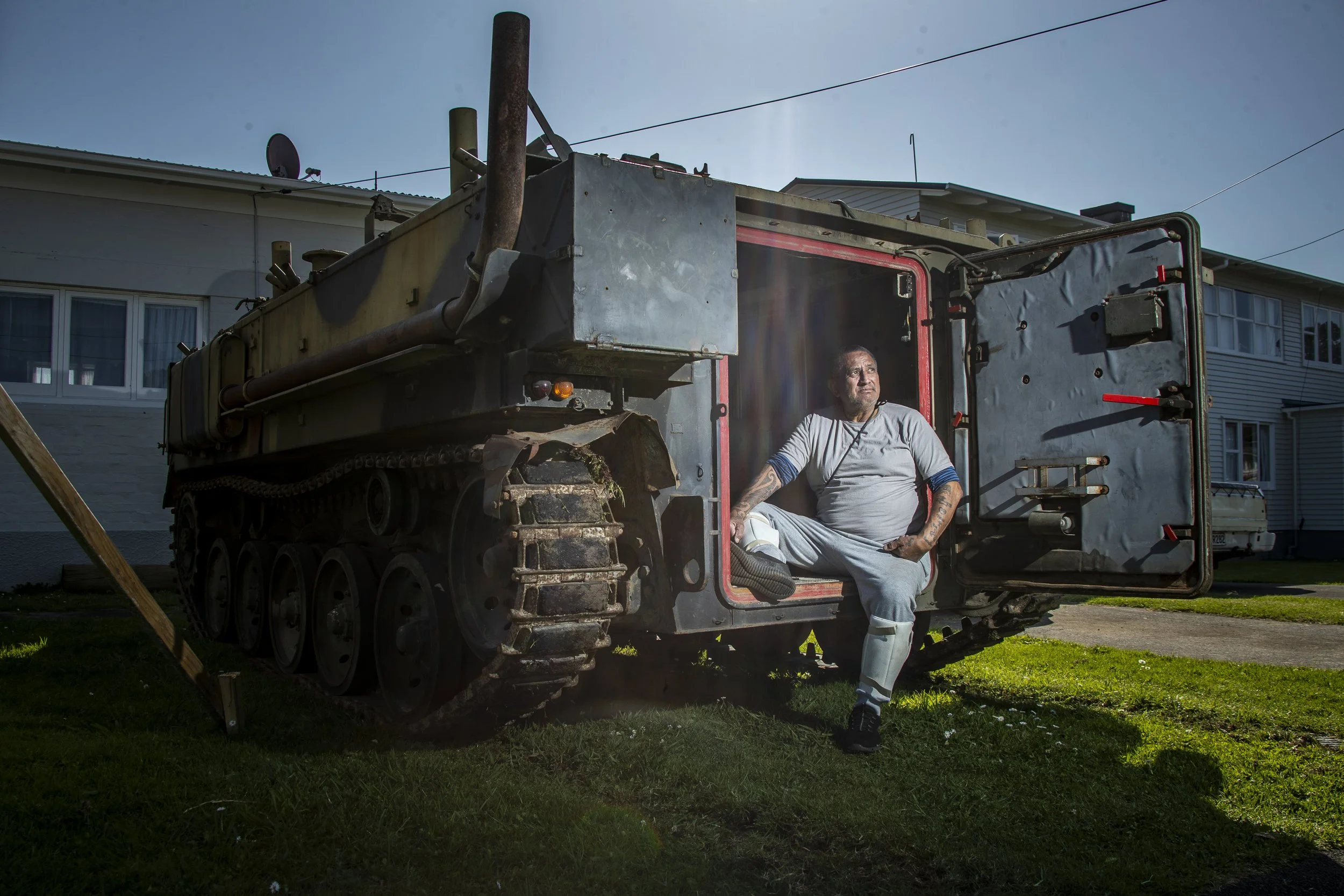 A man with tattoos sitting on the doorway of a damaged military tank in a residential area.