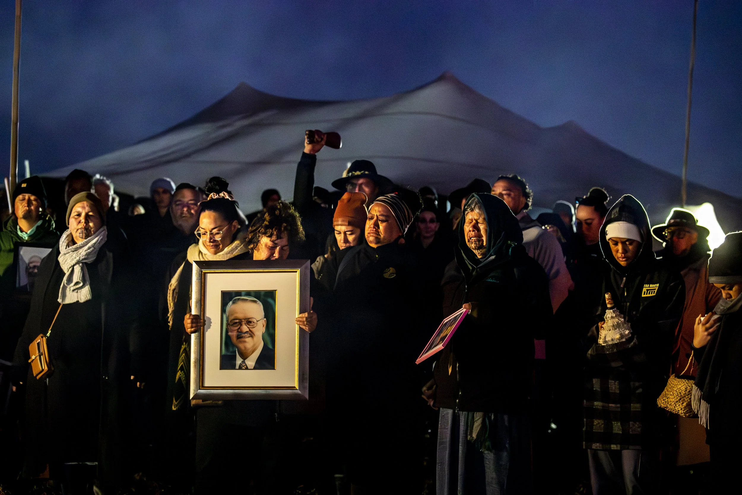 A group of people mourning at a vigil or memorial, some holding framed photographs of a man with glasses and a suit, others with serious or teary expressions, standing under a dark sky with a large mountain in the background.