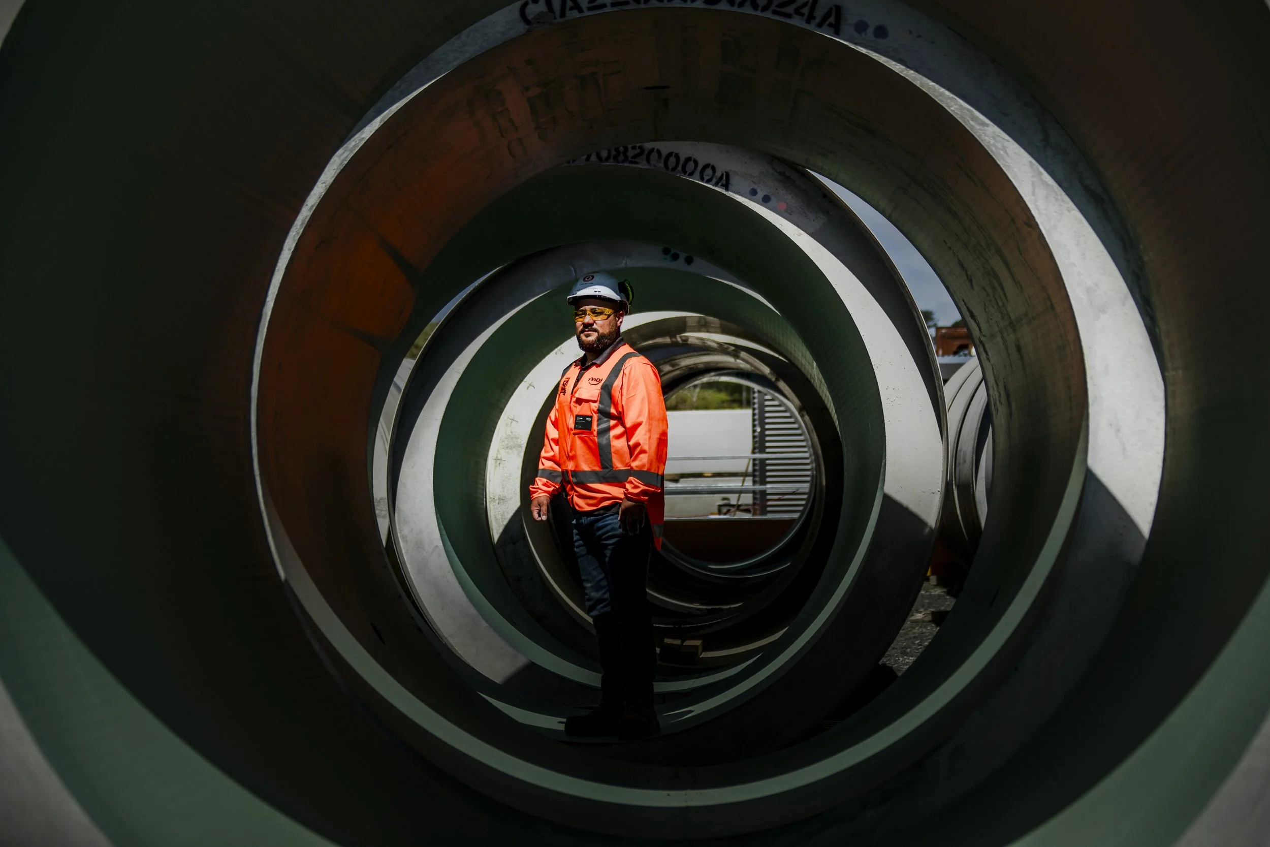 A construction worker in an orange safety jacket, black pants, a safety helmet, and sunglasses standing inside a large metallic pipe at a construction site.