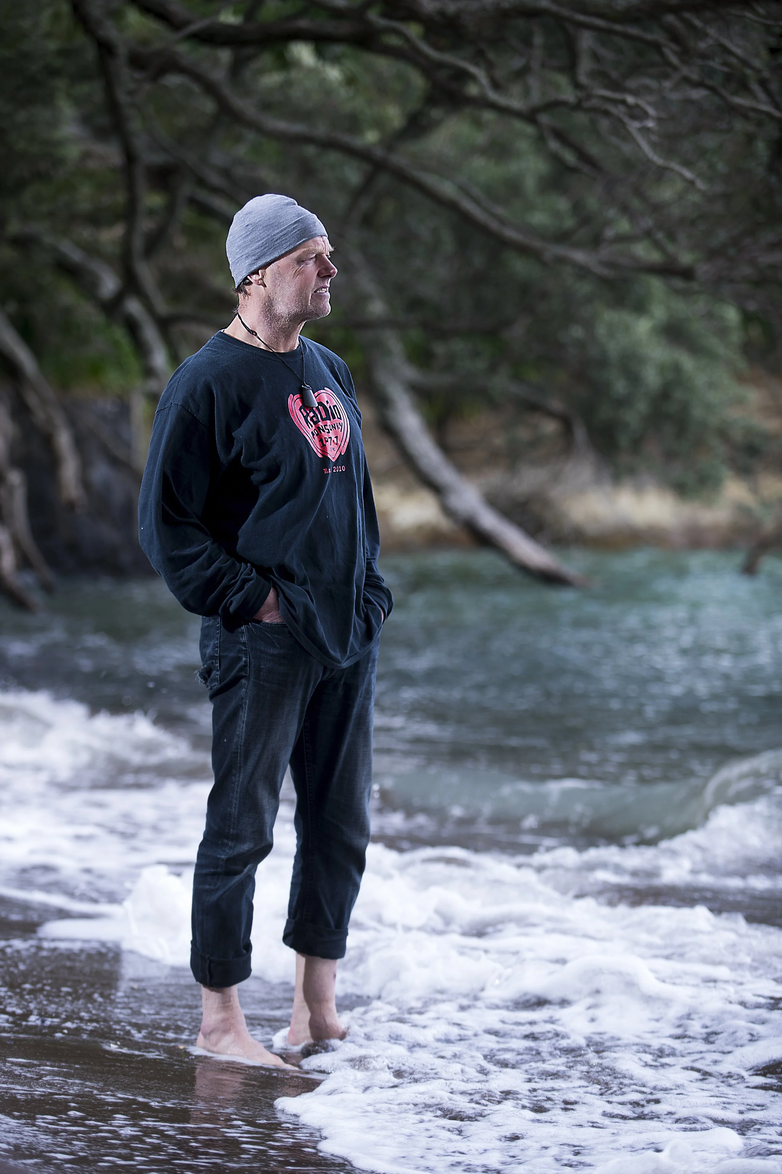 A man wearing a gray beanie, black sweatshirt, and rolled-up jeans stands barefoot in the ocean waves on a beach, with trees and rocky shoreline in the background.