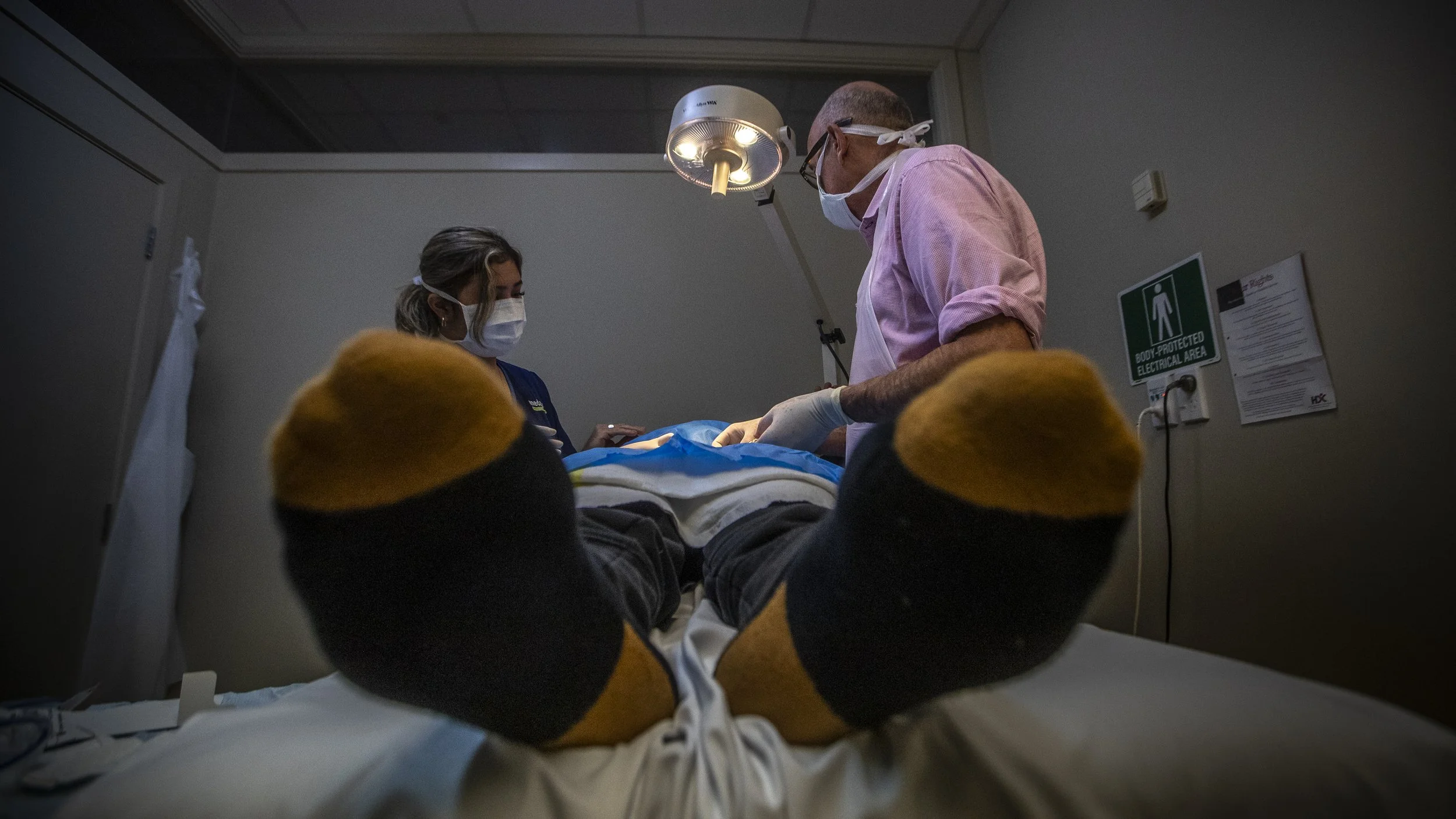 Medical professionals performing a procedure on a patient lying on a hospital bed, viewed from the patient's perspective at foot level.