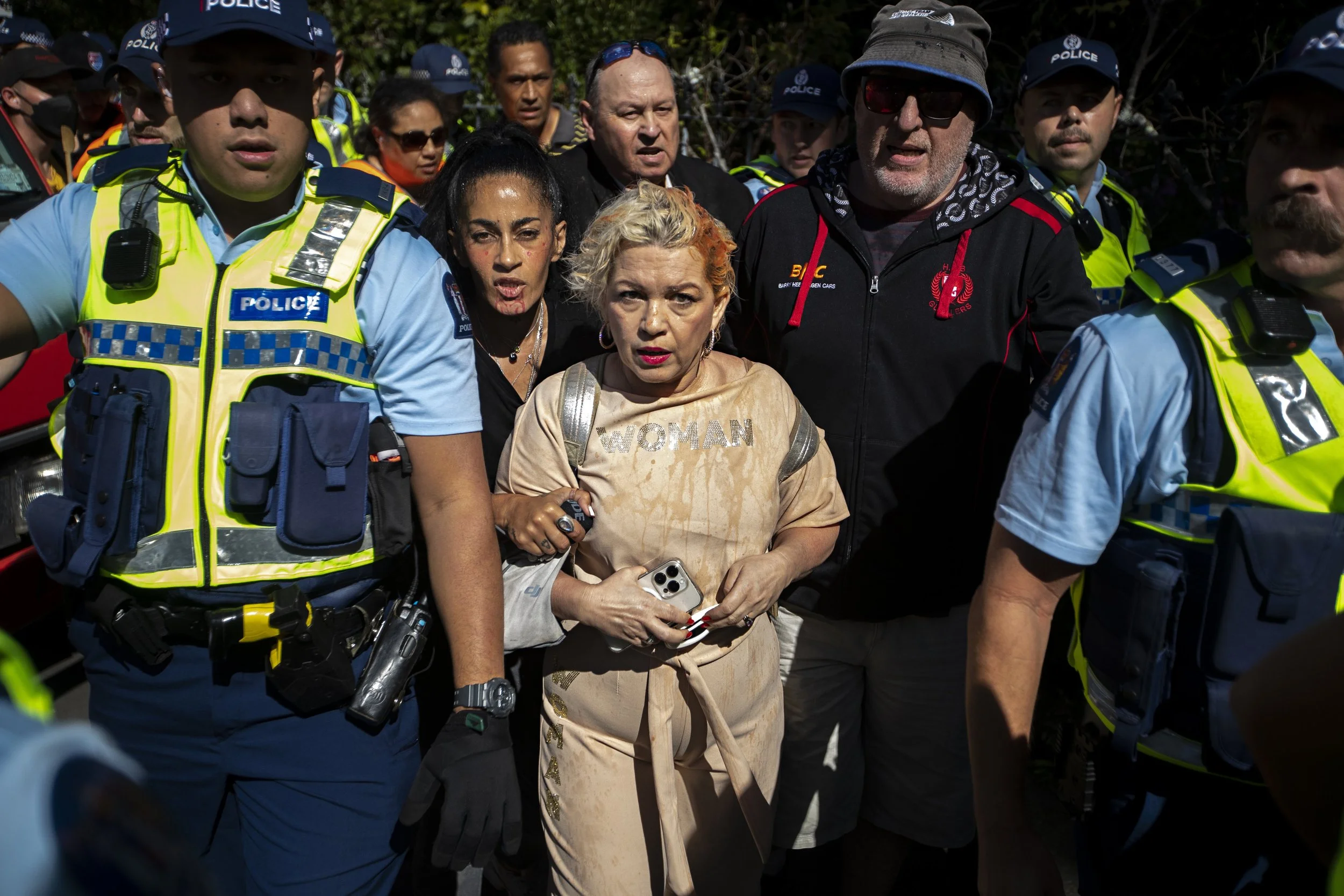 A group of police officers and civilians gather outdoors, with a woman in the center wearing a beige outfit with the word 'WOMAN' on her shirt, holding a phone and a small object, all looking serious amid a tense situation.