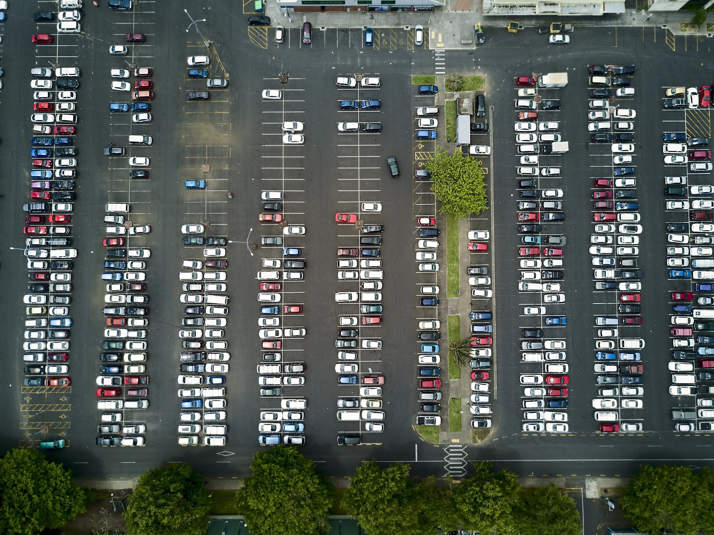 An aerial view of a parking lot filled with cars arranged in rows, with some trees lining the bottom and a small landscaped area with grass and shrubs in the center.
