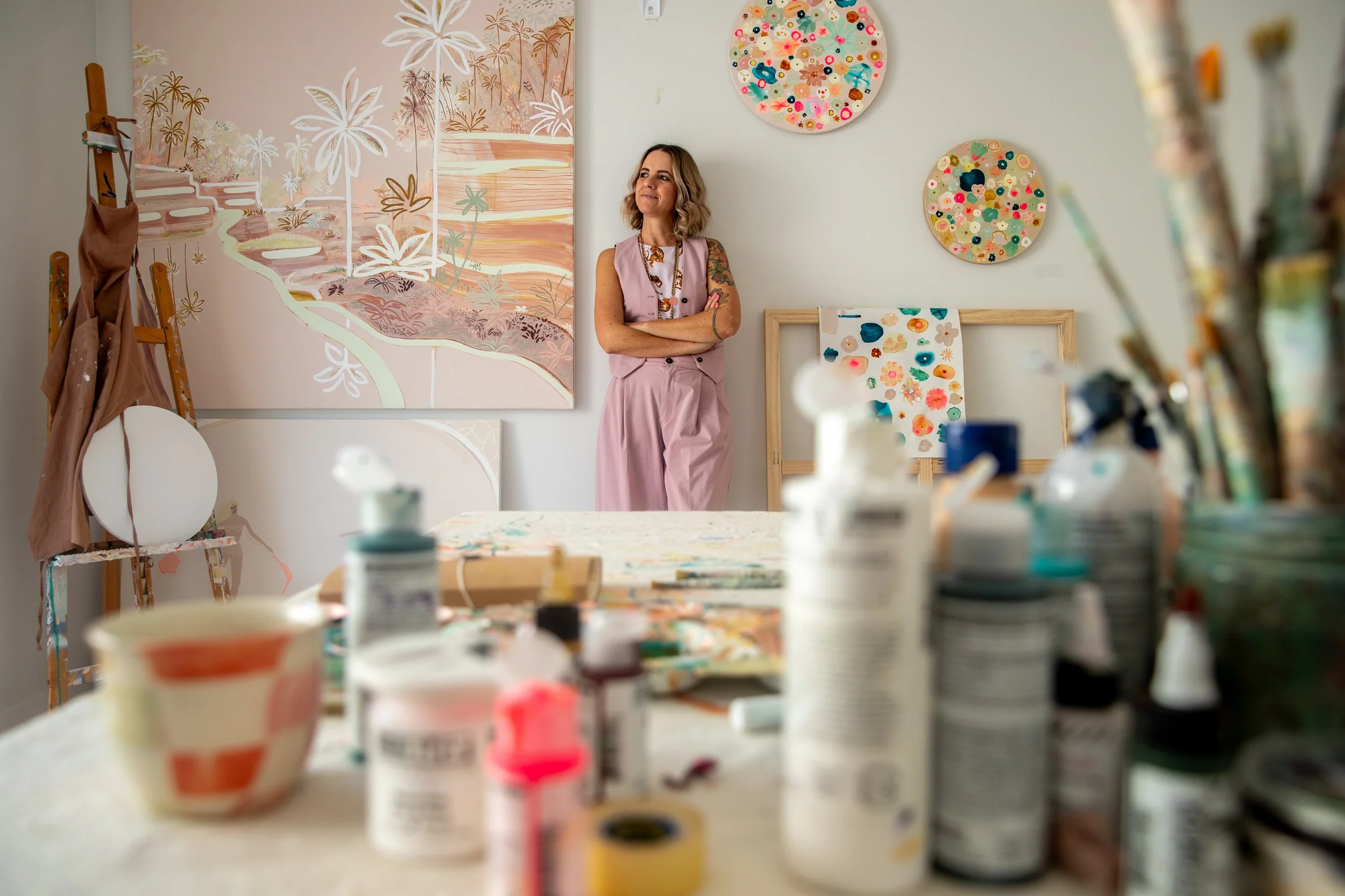 An artist's studio with a woman standing against a white wall decorated with colorful, circular art pieces. The studio has a large abstract painting of a landscape with trees and water, and numerous art supplies on a table in the foreground.
