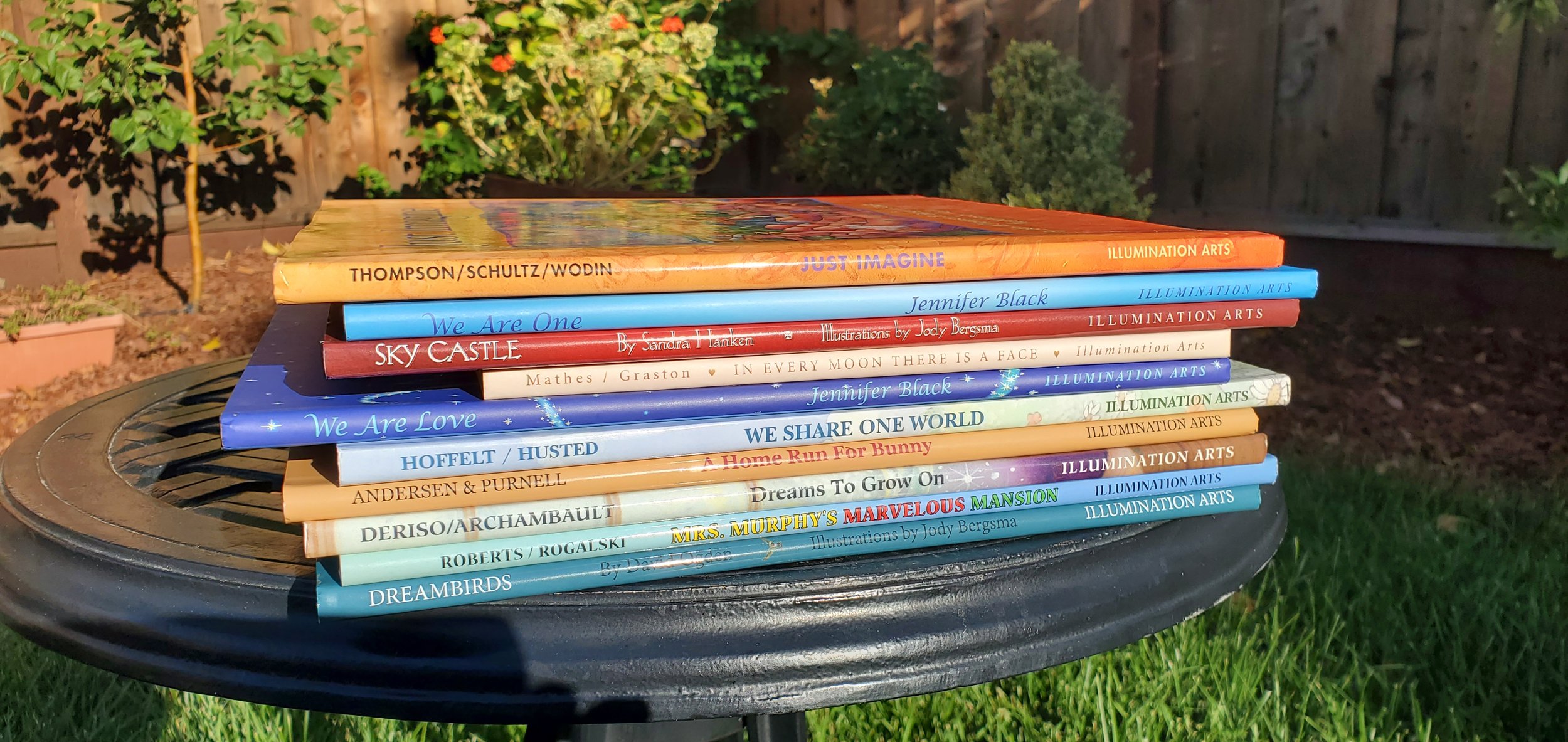 A stack of children's books on a round black outdoor table in a garden with plants and a wooden fence in the background.