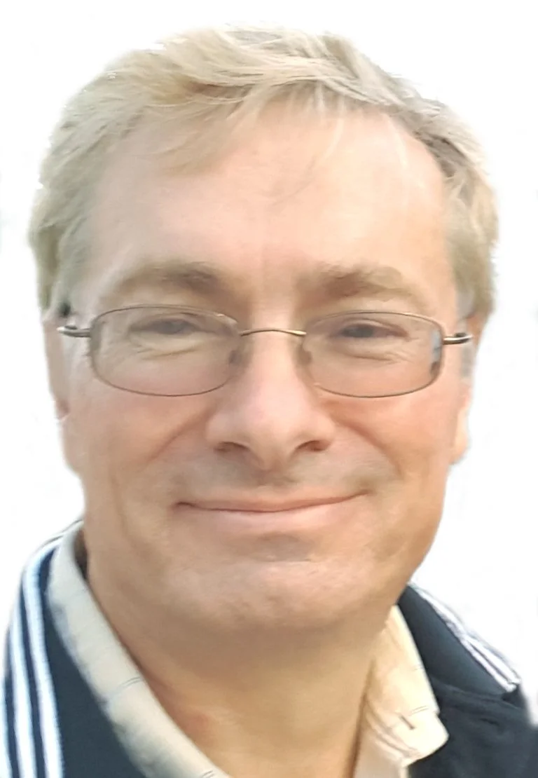 Close-up of a middle-aged man with glasses, light-colored hair, smiling, wearing a collared shirt and sweater.