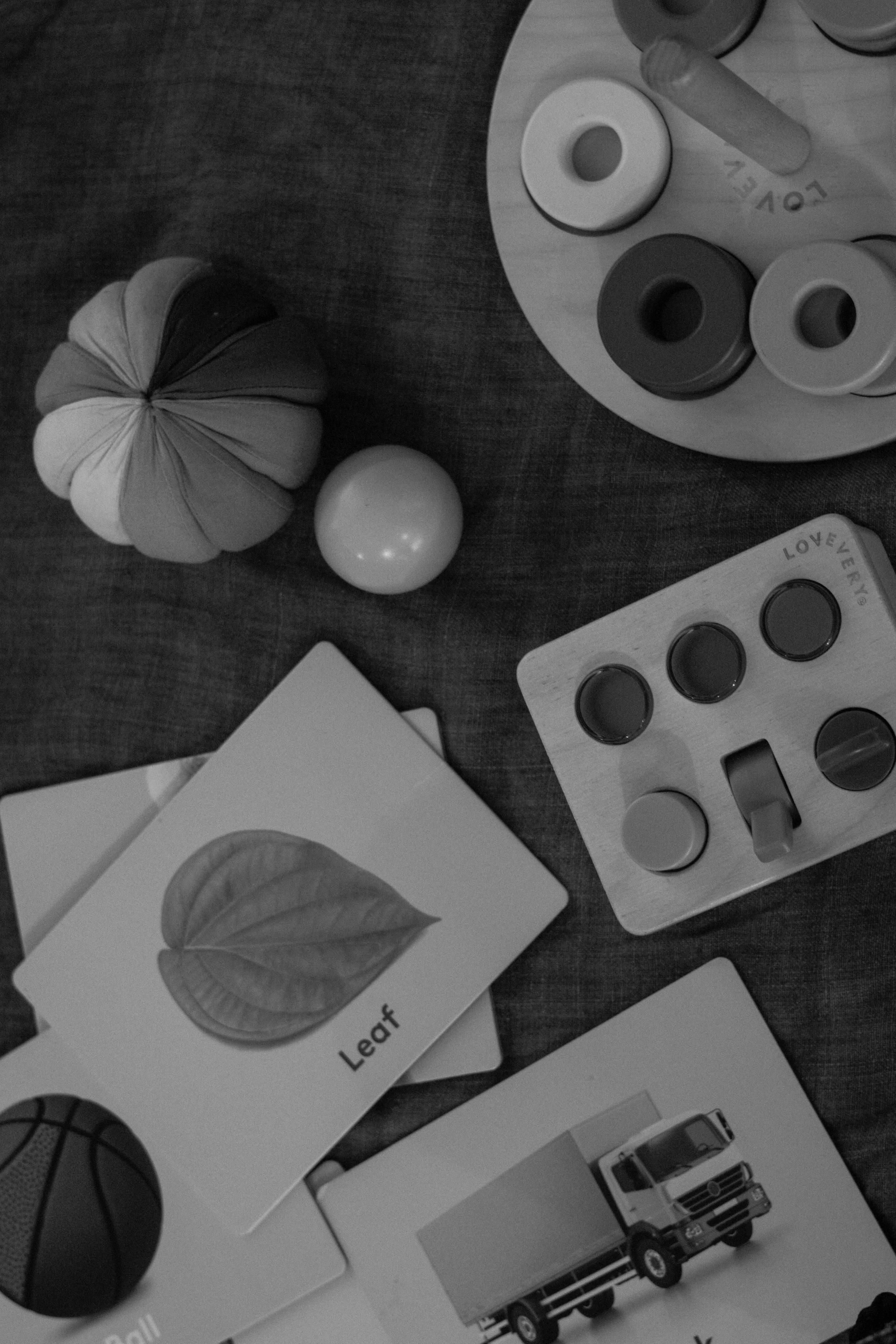 A collection of educational toys and cards on a table, including a pumpkin, a ball, a wooden box with colored rings, flashcards with images and words like "Leaf," and a toy truck.