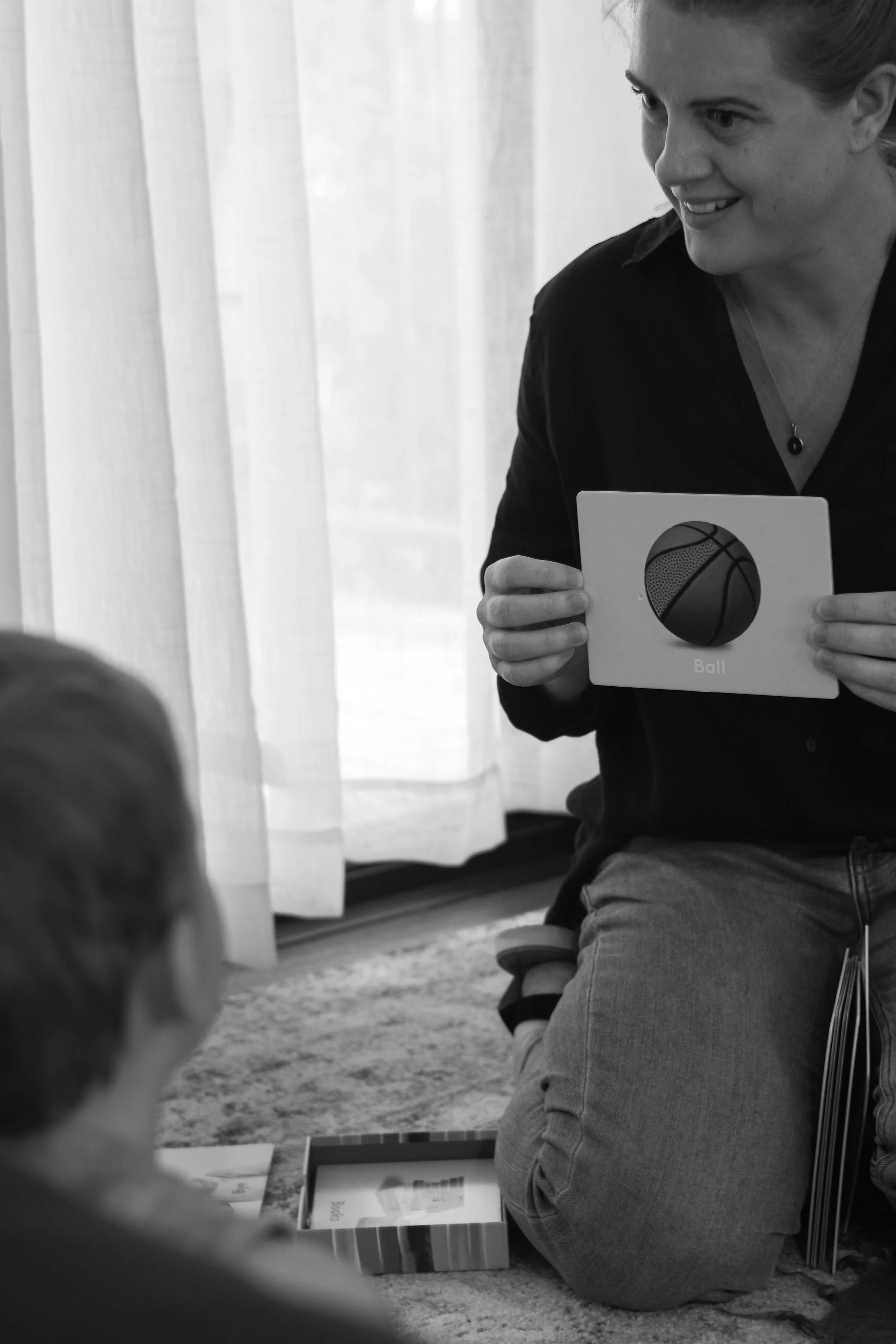 A woman holding a picture of a basketball and a ball toy, kneeling on the floor in front of a child with a box on the floor nearby.