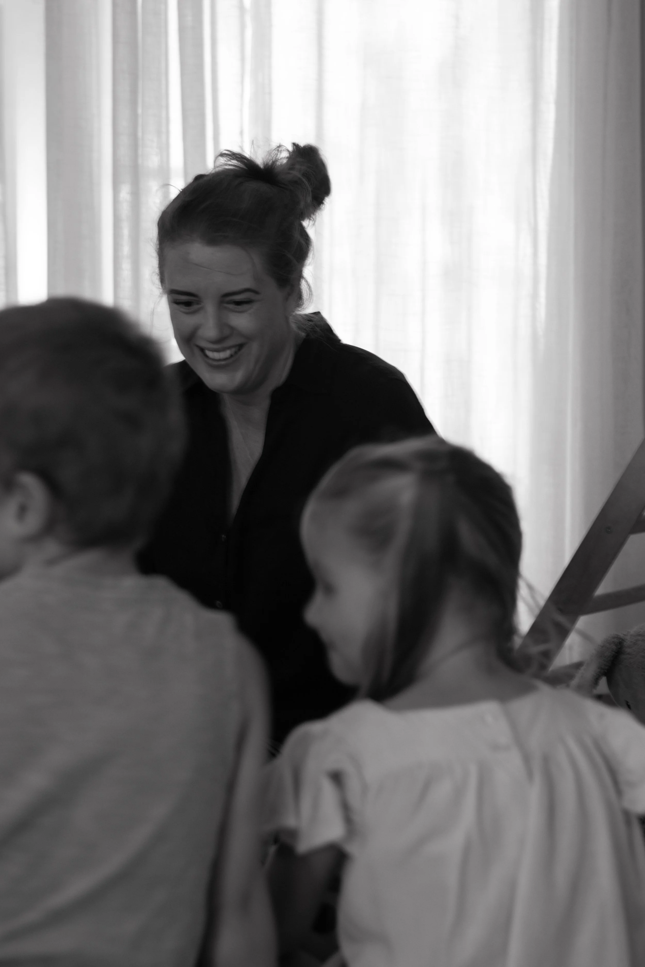 A woman with dark hair in a bun smiling and interacting with two children, a boy and a girl, in front of a light-colored curtain.