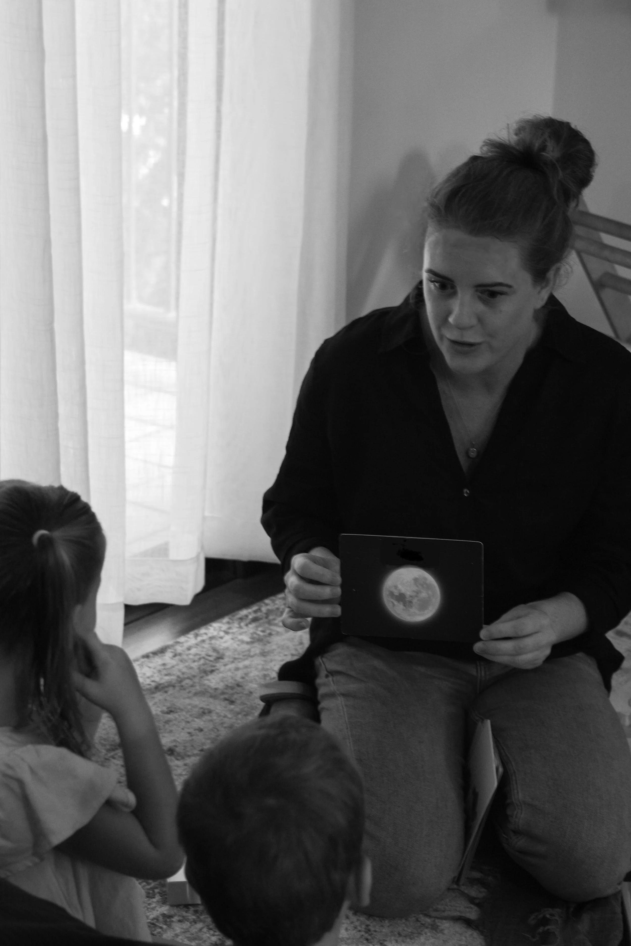 A woman sits on the floor holding a card with an image of the moon, teaching children about the moon in a cozy room with curtains and a rug.