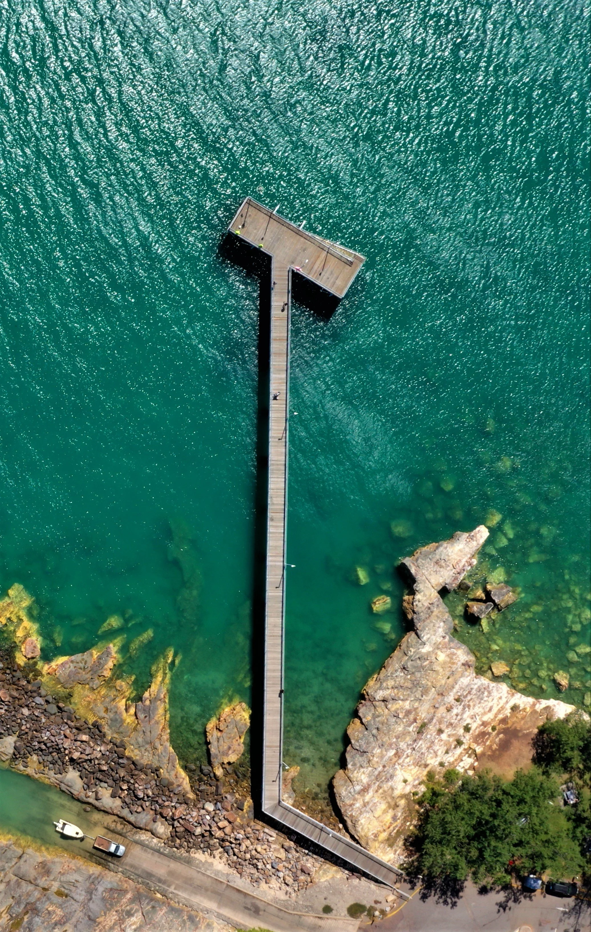Aerial view of a long wooden pier extending into clear green water, with rocky shoreline and trees nearby.