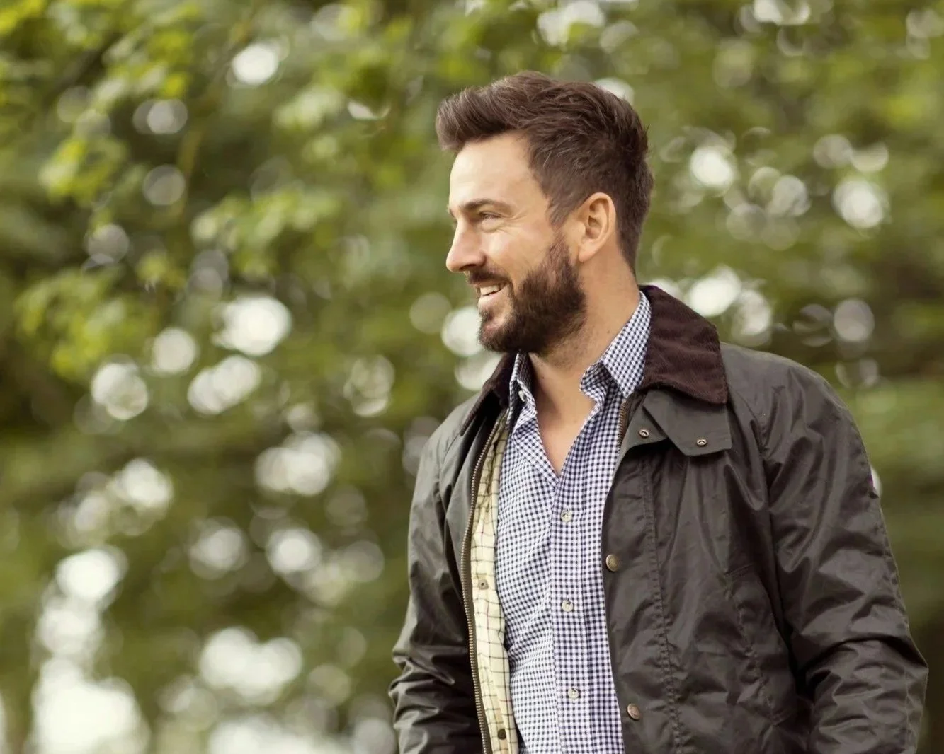 A man with a beard smiling outdoors, wearing a checkered shirt and a dark jacket, with a blurred green leafy background.