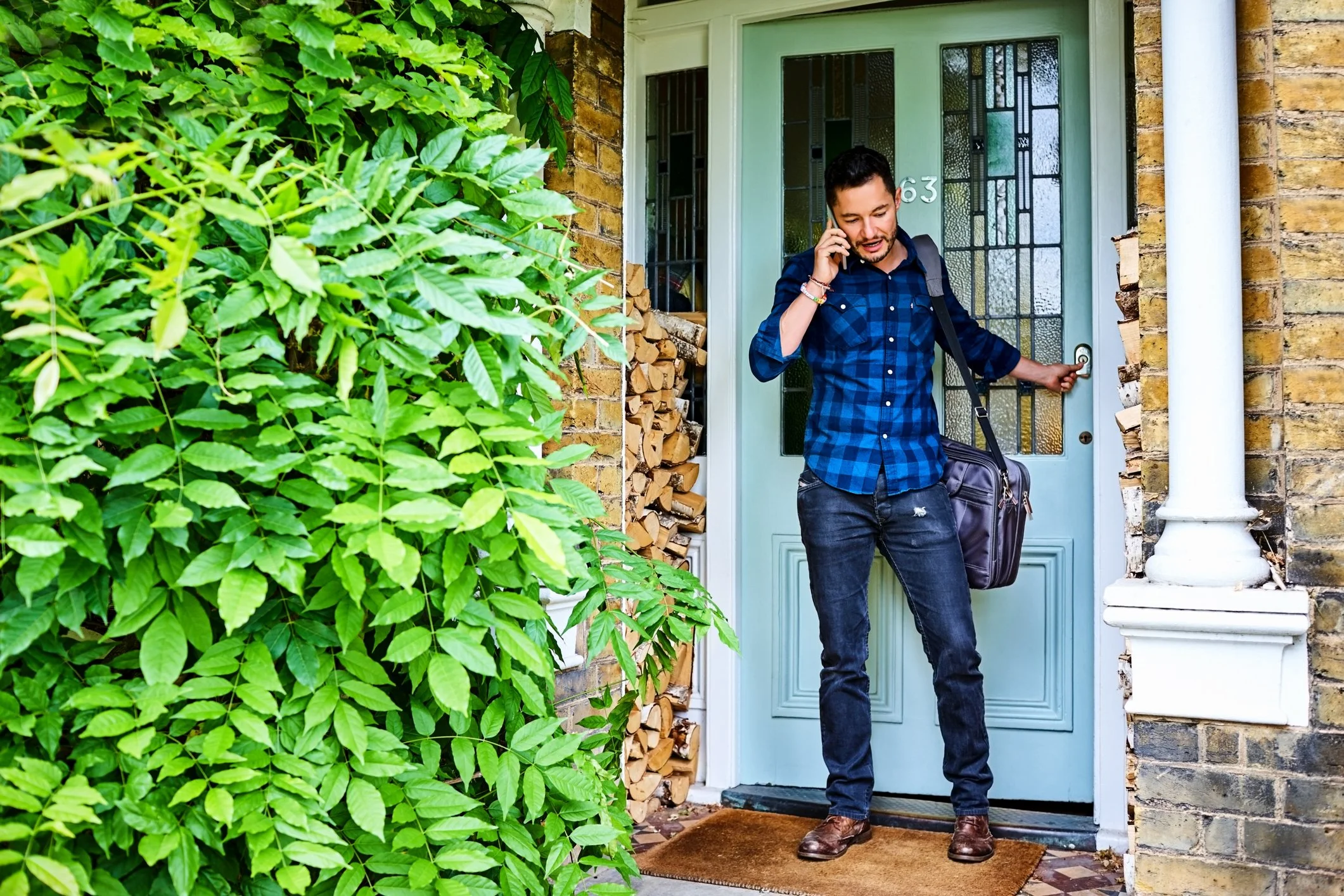 Man standing at the front door of a house, talking on cell phone, holding the door handle with one hand, carrying a black shoulder bag, dressed in a blue checked shirt, jeans, and brown shoes, with a stack of firewood beside the door, surrounded by green foliage.