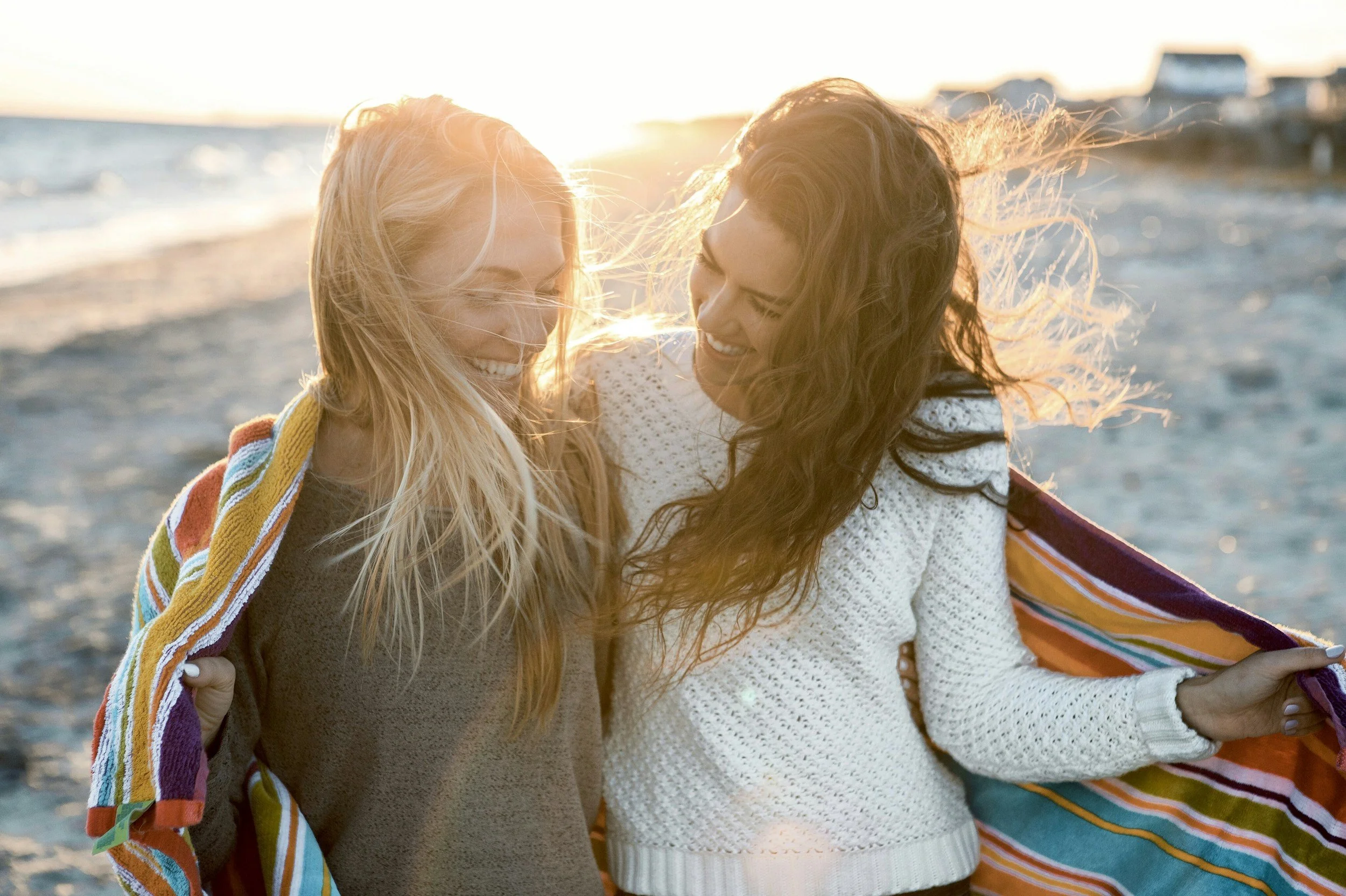 Two women at the beach holding a colorful towel, smiling, with the sun setting behind them.