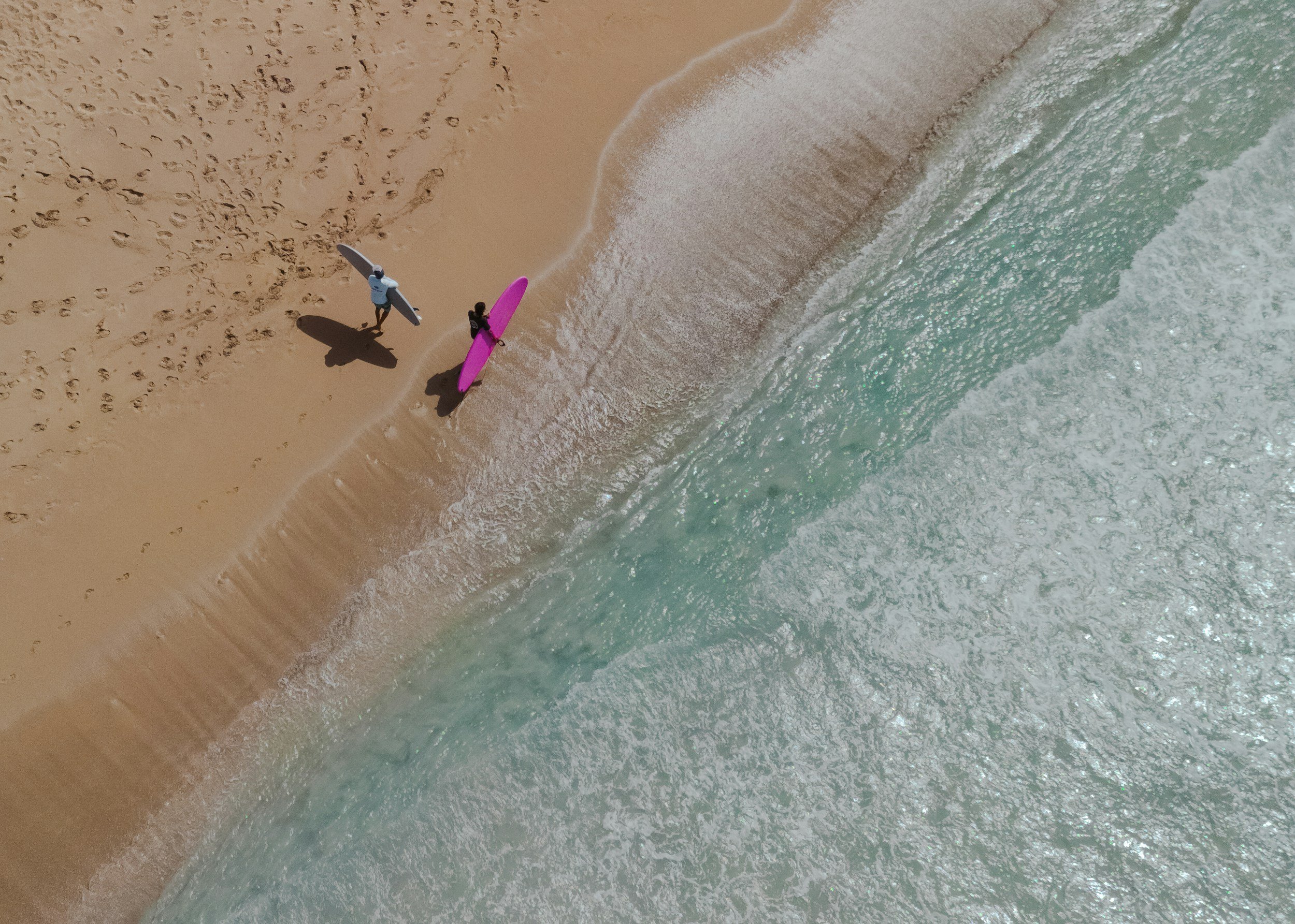 Aerial view of two surfers walking on the sandy beach with surfboards, one with a blue board and one with a pink board, near the shoreline with waves crashing.
