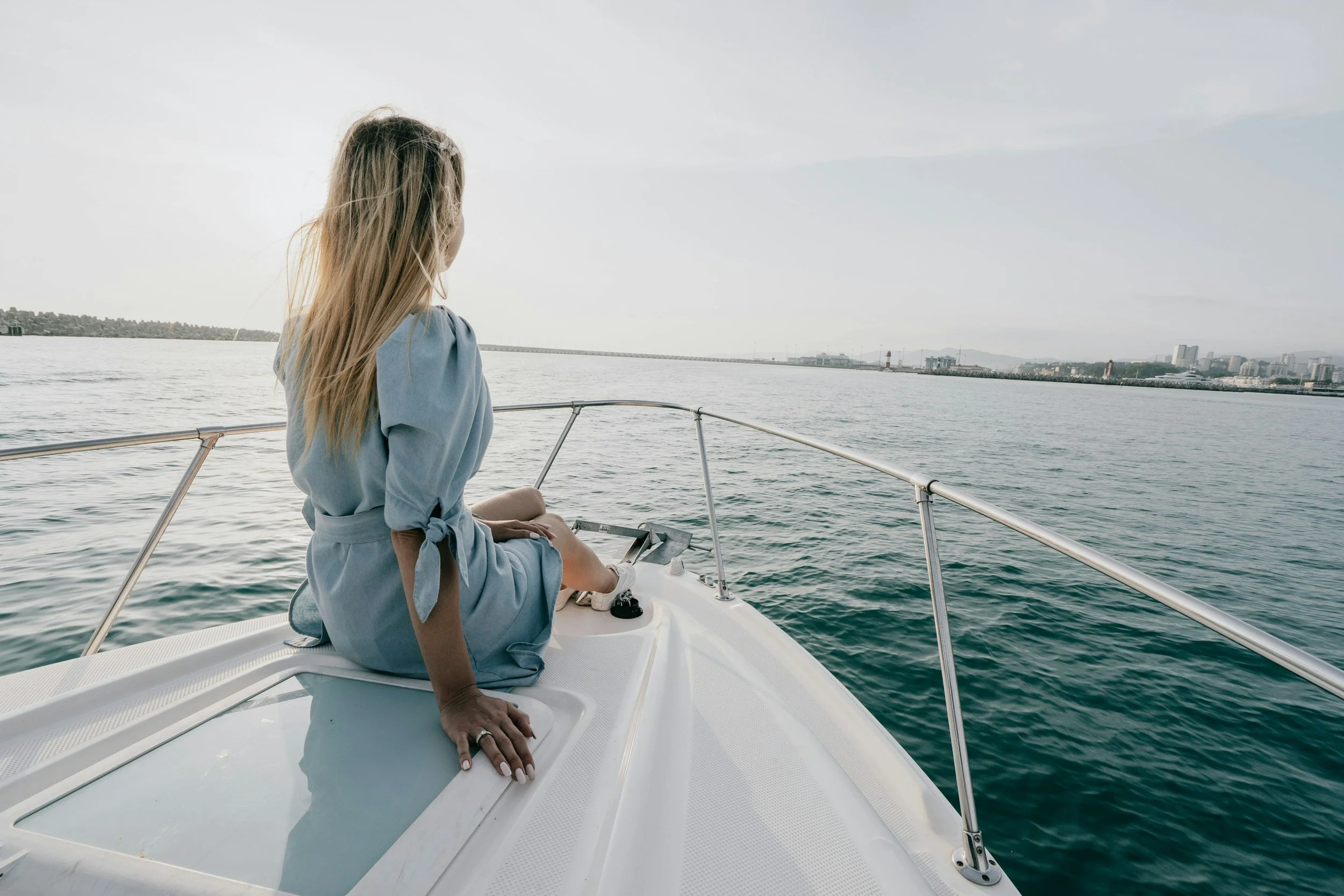 Woman in a blue dress sitting on the edge of a boat looking out at the water and city skyline in the distance