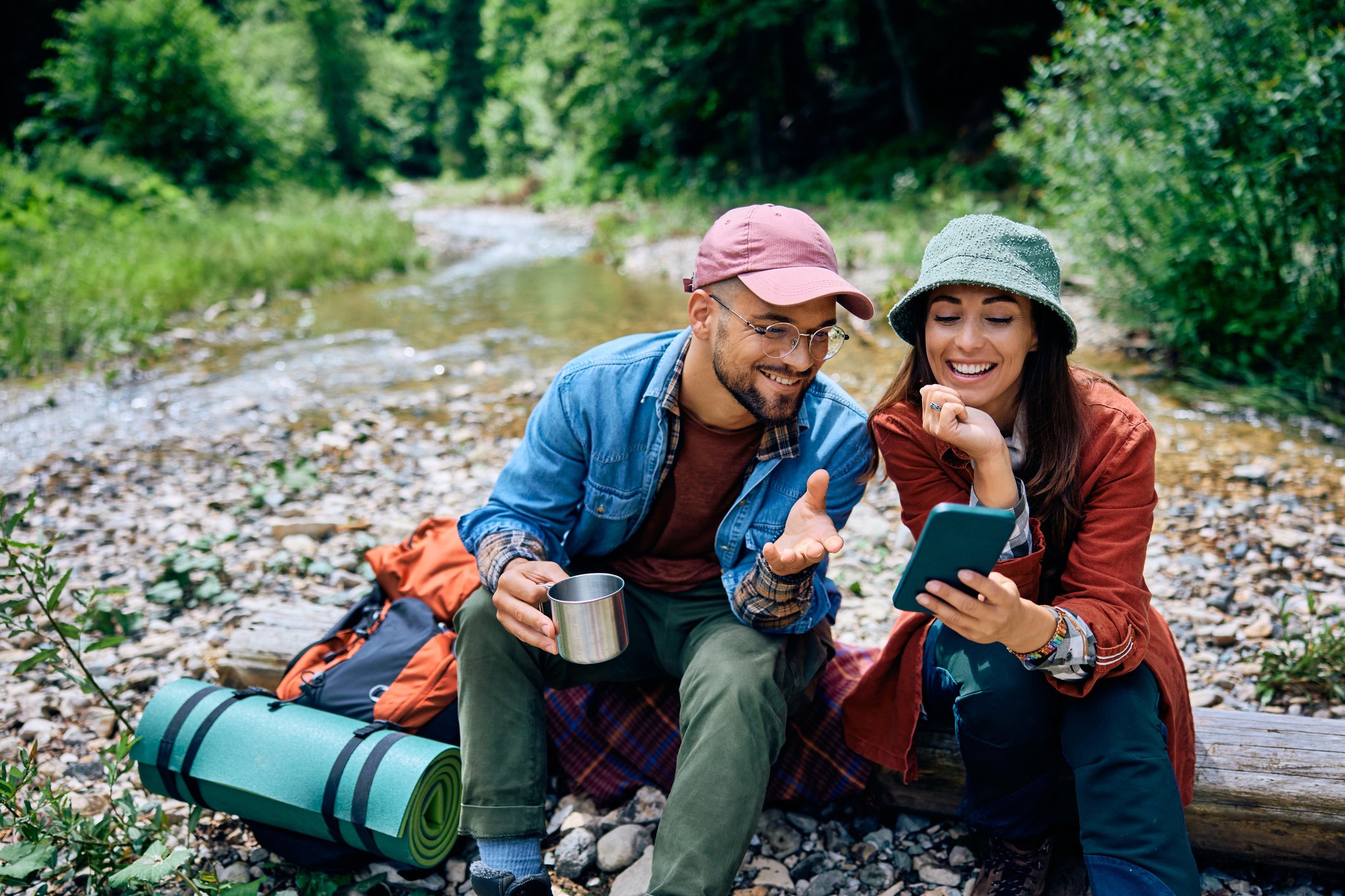 A smiling man and woman sitting on a log by a stream in a forest, looking at a smartphone together, with a backpack and a rolled-up sleeping pad nearby.