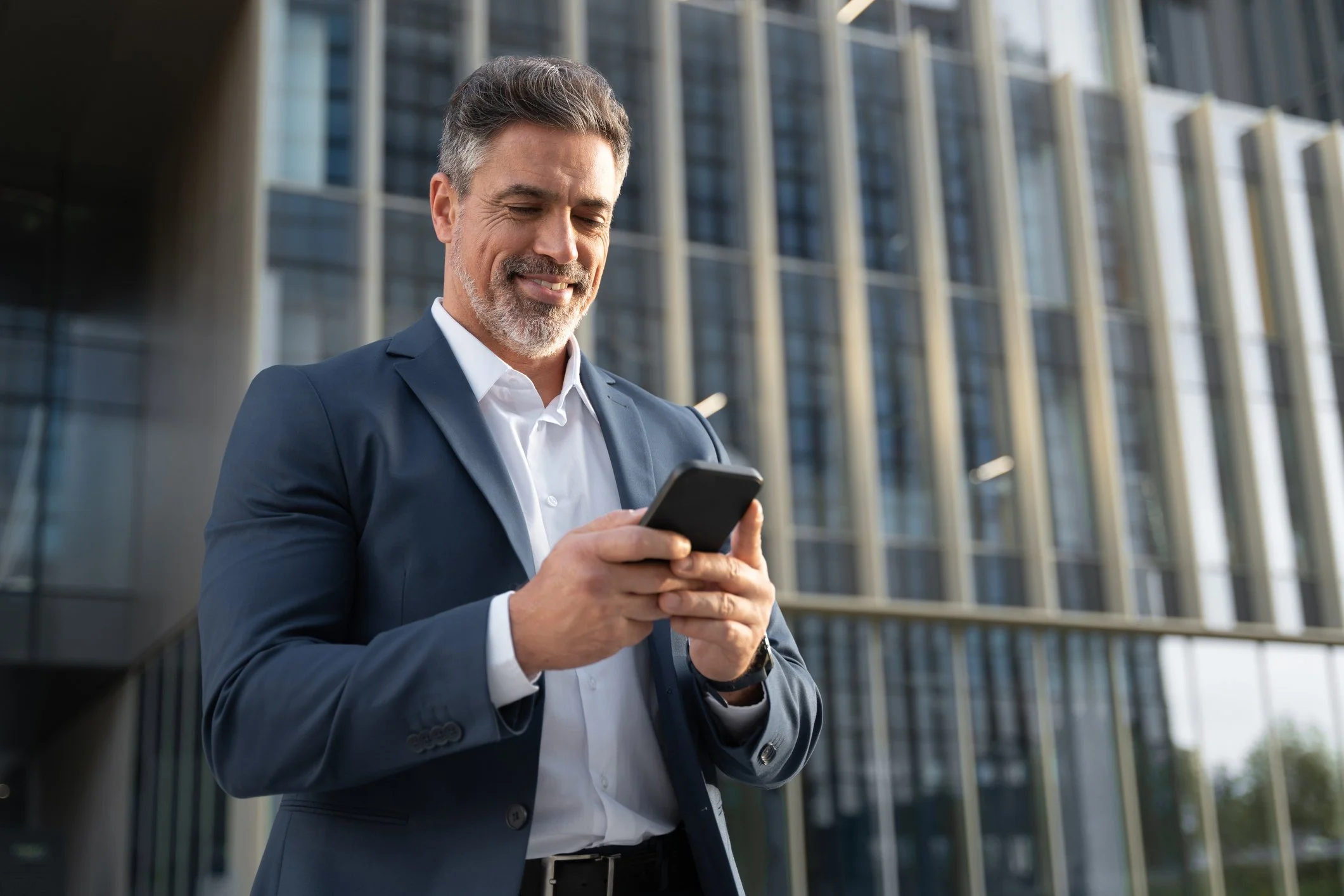 A middle-aged man in a business suit smiling while looking at his smartphone outside a modern office building.
