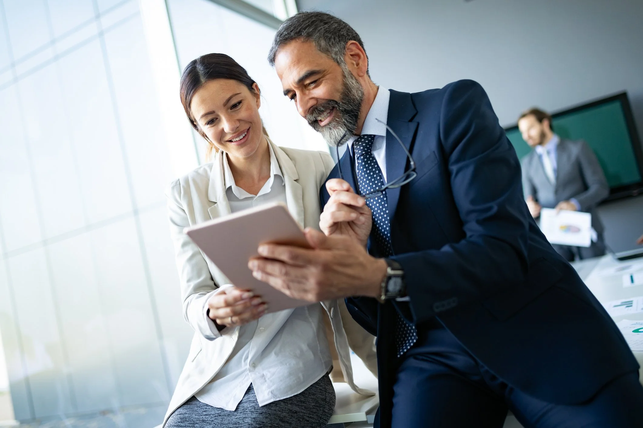 Two business professionals, a woman and a man, looking at a tablet together in an office, with another man in the background at a conference table.