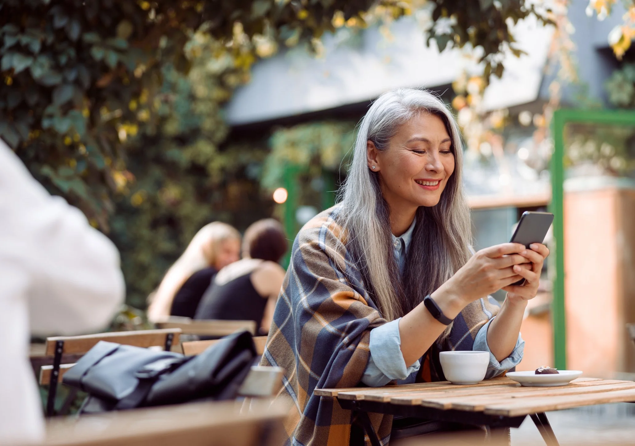 Smiling woman with gray hair sitting at an outdoor cafe table, looking at her phone, with a bowl and a plate of food in front of her. Other people are blurred in the background.